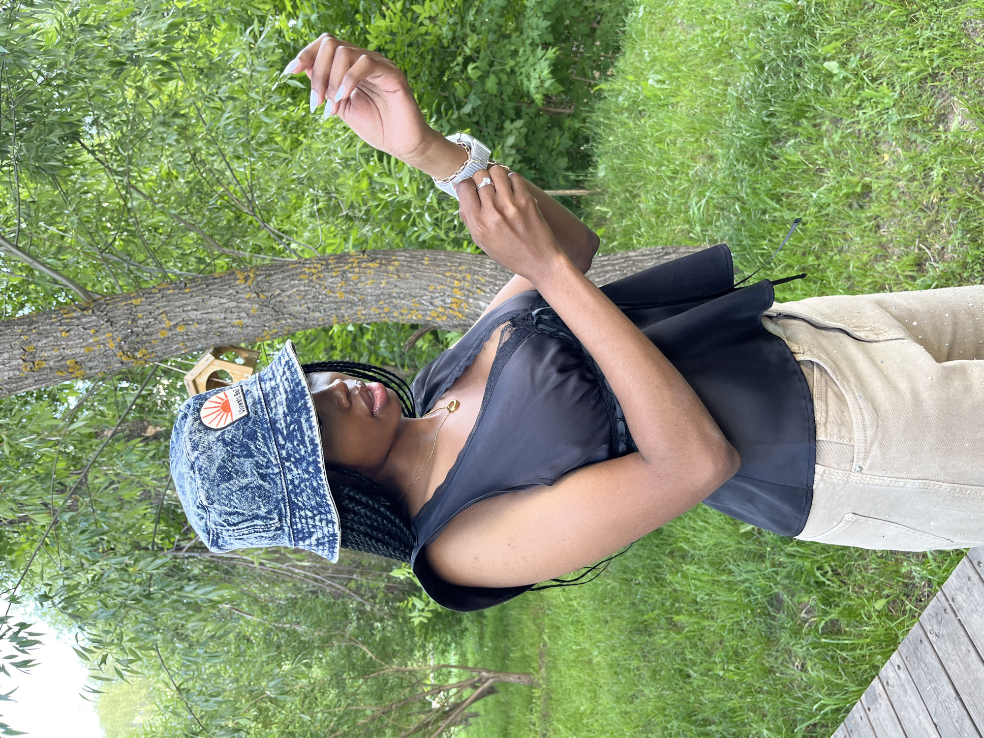 A young Black woman stands outdoors in what appears to be a park, possibly in Kazan, Russia, given the context. She is wearing a blue acid-wash denim bucket hat with a patch featuring a rising sun and text. She has on a black sleeveless top with lace detailing and a ruffled neckline, paired with light-colored pants adorned with small rhinestones. Around her neck is a delicate gold necklace. Her right hand is raised, and she is adjusting a watch on her left wrist, which also features a bracelet. The background consists of lush green grass, trees with textured bark, and foliage, suggesting a natural, possibly wooded, setting. A wooden structure, possibly a birdhouse, is attached to a tree trunk behind her. The lighting indicates it is daytime, with soft shadows and a bright sky, though the sky itself is not directly visible in the frame.