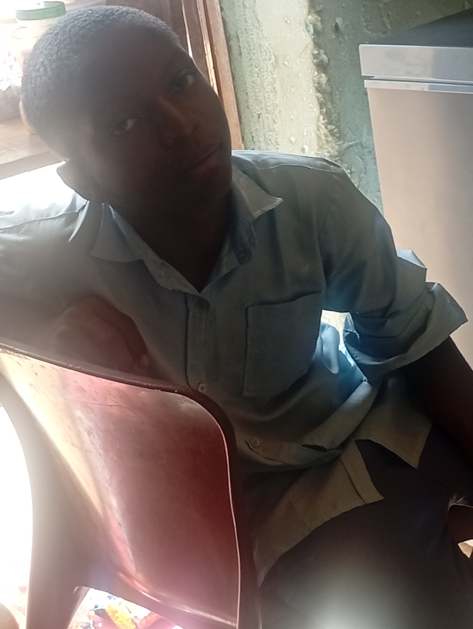 A young Black man is seated on a red chair inside a building in an unknown location. He is wearing a light blue collared shirt with a pocket. A white refrigerator is visible in the background. The wall behind him is painted in a distressed, light-colored pattern.