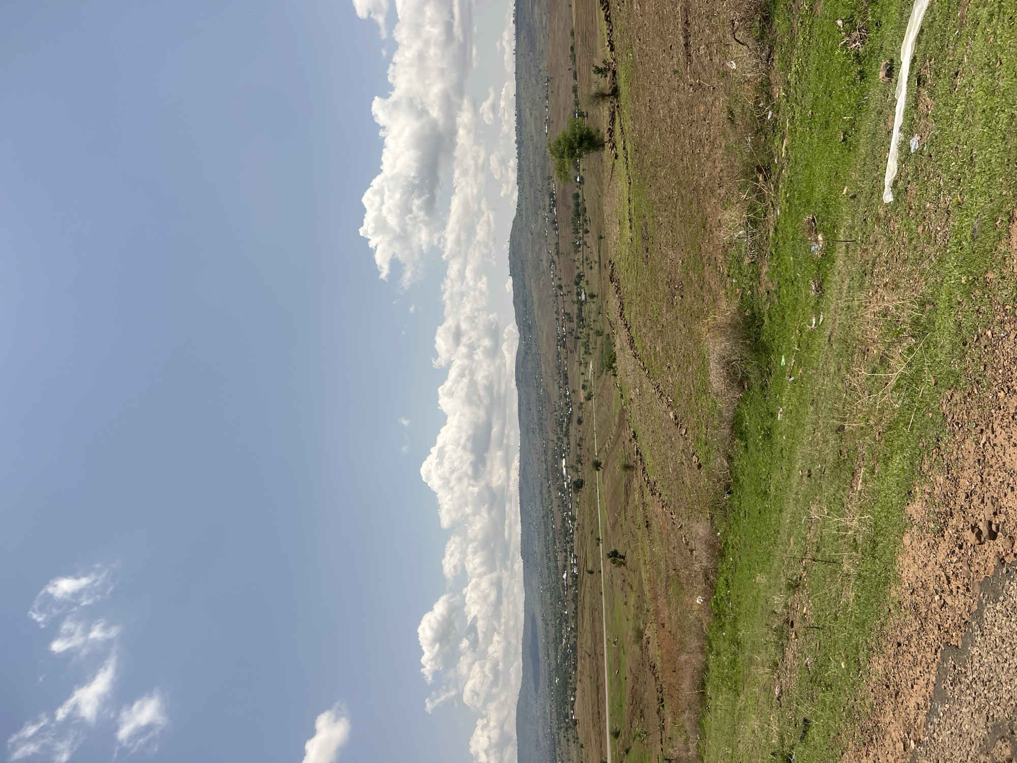 The image depicts a wide-angle landscape under a partly cloudy sky, characteristic of a rural or semi-urban environment in Chum, Nigeria. The upper half of the frame is dominated by a light blue sky populated with various cumulus clouds, ranging from wispy formations to larger, more defined clusters.
The horizon line reveals a series of undulating hills or elevated terrain stretching across the entire width of the background. Below these hills, numerous low-rise structures form scattered settlements or villages, indicative of human habitation, distributed across the midground.
A two-lane paved road, likely asphalt, traverses horizontally through the midground, exhibiting a gentle curve. Agricultural fields and open land flank both sides of this road. The vegetation in these fields is varied, presenting dry, brownish patches of soil alongside areas of greener growth. Sections of the fields show visible stone lines or low walls, potentially indicative of terracing or property demarcation. A few isolated trees with green foliage are sparsely distributed across the midground.
The immediate foreground consists of an unpaved area on the left with visible dirt, gravel, and small rocks, transitioning into an uneven expanse of mixed green grass and dry, straw-like vegetation. Anthropogenic debris is present in the foreground, including a distinct long piece of white plastic sheeting or fabric on the lower right and several smaller fragments of white litter scattered among the grassy areas. No individuals, animals, or ongoing activities are visible within the frame.
