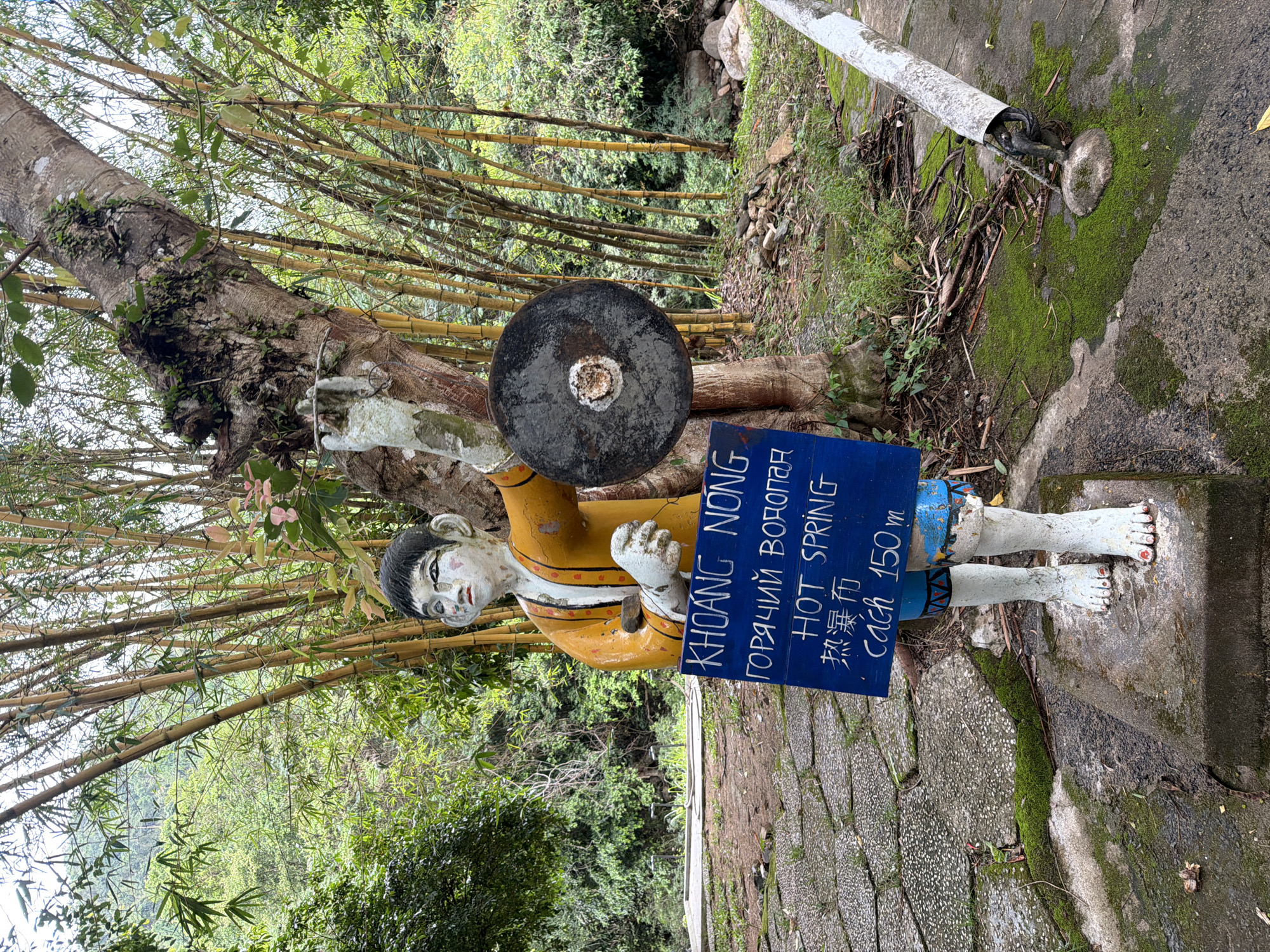 A weathered statue of a boy in a yellow tunic stands in front of a cluster of bamboo stalks and a large tree. The statue holds a circular object in its right hand. A blue sign with white lettering is positioned in front of the statue. The sign contains text in multiple languages. The first line, "KHOANG NONG," is in blue, followed by "горячий водопад" in white Cyrillic script. Below that, "HOT SPRING" is written in white. Further down, "熱瀑布" appears in Chinese characters, and the last line reads "cách 150 m" in blue. The ground around the statue is paved with stones and covered in patches of green moss. In the background, lush green foliage and trees are visible. To the right of the statue, a weathered white pipe emerges from the ground. The scene suggests an outdoor location, possibly a natural attraction. This image is of the Khoang Nong Hot Spring, located in Khanh Vinh City, Vietnam.