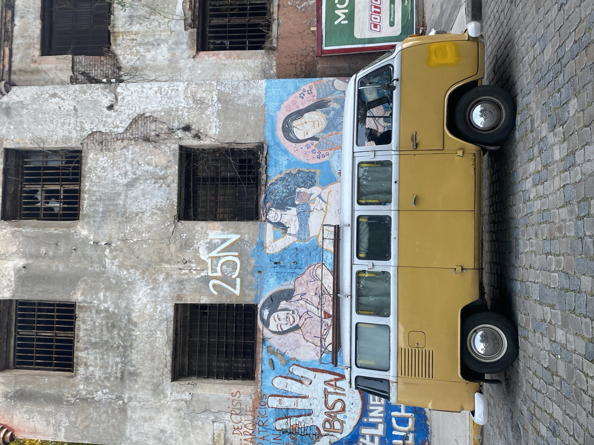A yellow and white Volkswagen van is parked on a cobblestone street next to a decaying building in Buenos Aires, Argentina. The building facade has multiple barred windows and is adorned with street art. The street art features images of faces and handprints, alongside the words "BASTA," "25N," and other phrases in Spanish, including the partial phrase, "DE CRISIS A TRICIOS PARQUE." The building's plaster is cracked and peeling, revealing the underlying brickwork in places. A green and red sign is partially visible on the upper right of the frame, positioned above the van. The van's windows are partially reflective, hinting at the presence of someone inside.