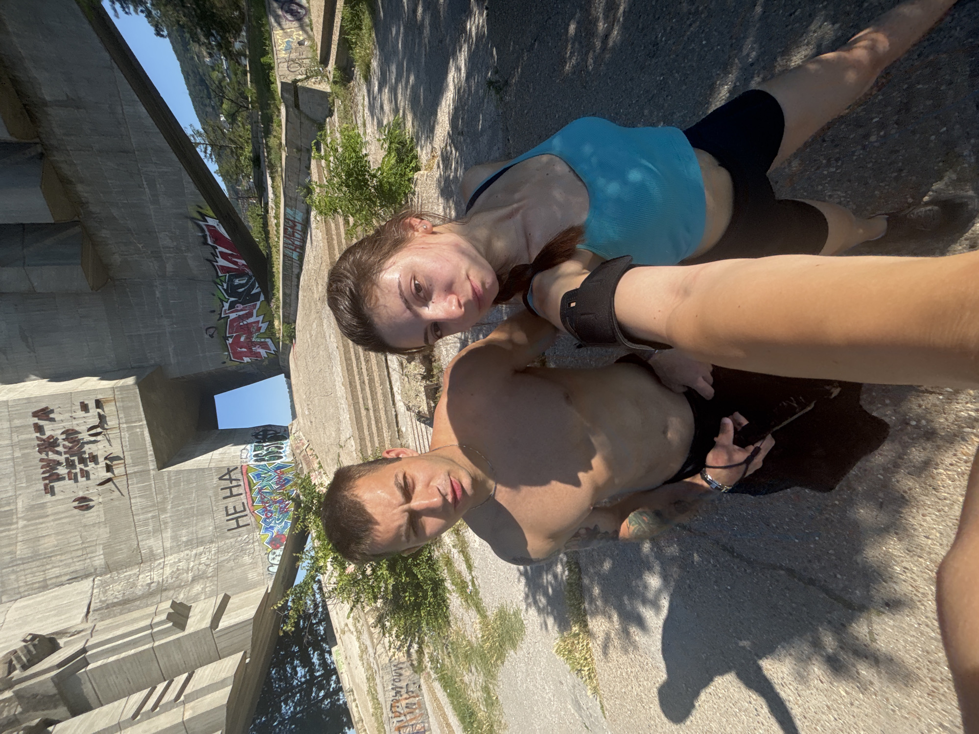 The image depicts a selfie taken by a young woman in front of a large concrete monument with graffiti. The monument appears to be the Buzludzha Monument, located in the Stara Planina mountains, near the city of Varna, Bulgaria. The woman is wearing a blue tank top and black shorts, and her hair is braided. A man, shirtless, stands next to her, also looking at the camera. Both appear to be in athletic attire, suggesting they might be exercising. The background shows the imposing structure of the monument, the surrounding landscape, and more graffiti. The lighting suggests it is daytime, likely late morning or early afternoon, with bright sunlight casting shadows. The overall atmosphere is one of urban exploration or a visit to a historical site.