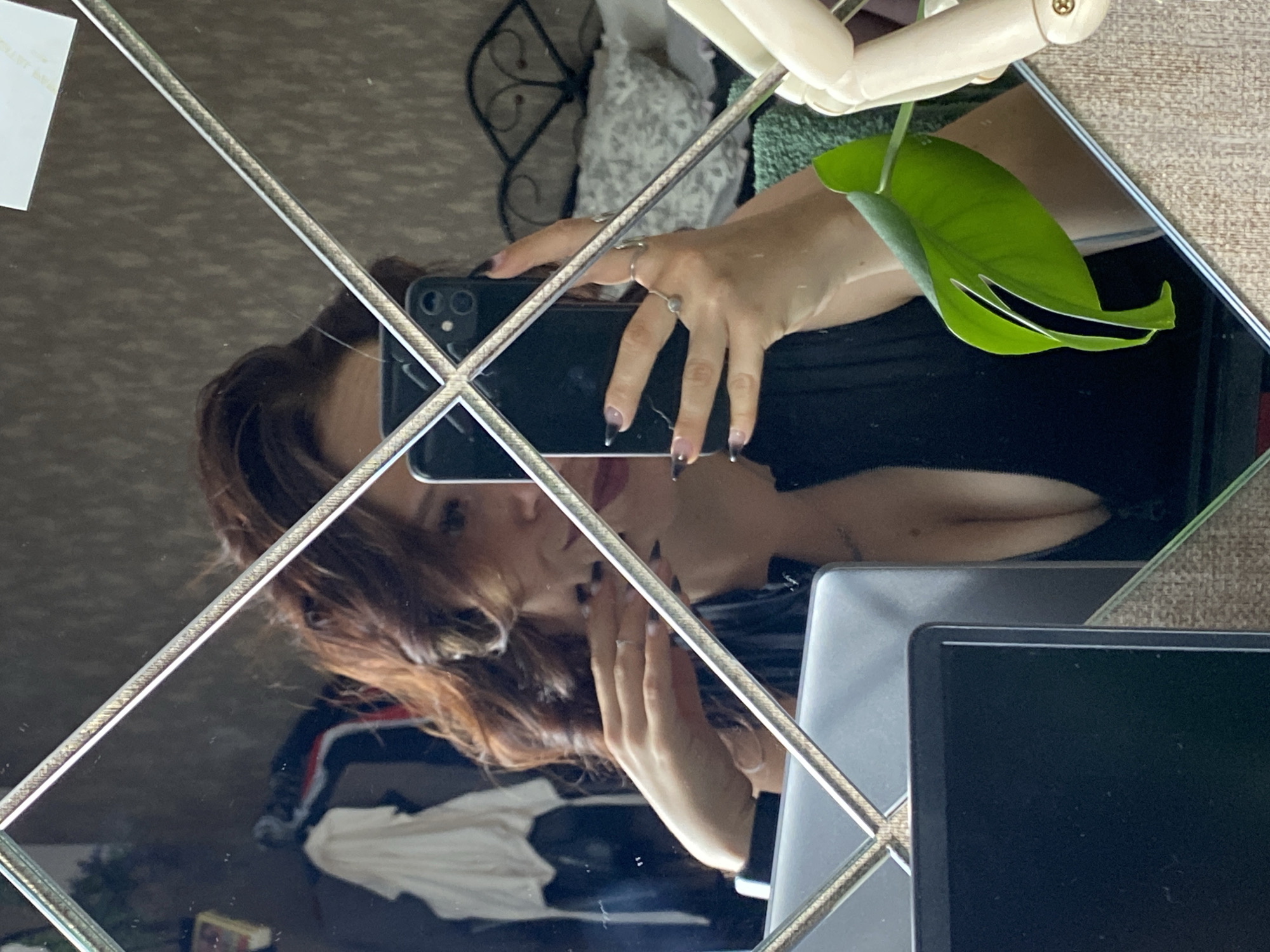 A mirror selfie taken by a person with long, reddish-brown hair. The person is wearing a black top and several rings on their fingers. Their fingernails are painted black. The reflection shows the person holding a black smartphone and touching their face. In the background of the reflection, there is a green leaf hanging from a wooden mannequin hand, and a glimpse of clothing and furniture. The mirror is framed by metallic-colored border creating a diamond pattern. The lighting suggests daytime.