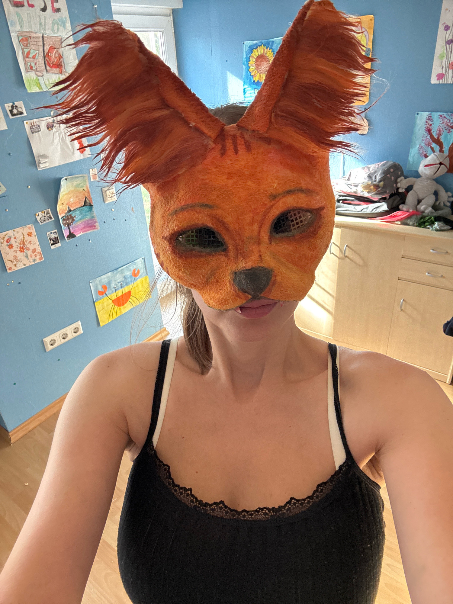 The image is a selfie taken indoors by a person wearing an orange fox mask. The mask has large, pointed ears with reddish-brown fur that appears windblown. The mask has mesh over the eye openings and a small black nose. The person is wearing a black tank top with white straps and black lace trim. The background consists of a light blue wall adorned with various drawings and pictures. To the right, there is a light-colored wooden cabinet with silver handles. The lighting suggests it is daytime, with sunlight casting shadows. The location is identified as Überherrn, Germany.