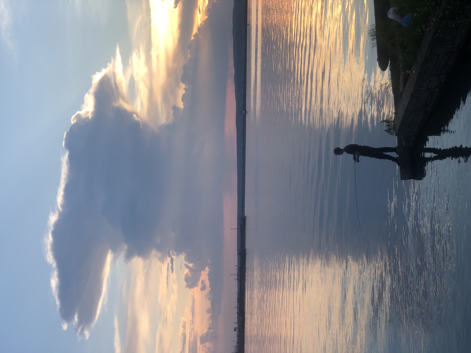 The image depicts a serene scene at sunset over a body of water, likely a river or reservoir, in Chaikovsky, Russia. The sky is a canvas of soft blues, oranges, and purples, with a dramatic, mushroom-shaped cloud formation dominating the upper left quadrant. The water's surface is calm, reflecting the colors of the sky and creating a mirrored effect.
In the foreground, a lone figure, silhouetted against the bright sky and water, is fishing from a concrete embankment. The person is standing, holding a fishing rod, and appears to be casting or reeling. The embankment is rough and appears weathered, with some vegetation growing along its edges. To the right of the fisherman, there are some discarded items, including a blue plastic bag and a bottle.
In the distance, a long, dark line of the horizon is visible, with what appear to be industrial structures and power lines dotting the landscape. The overall atmosphere is tranquil and evokes a sense of peace and solitude.