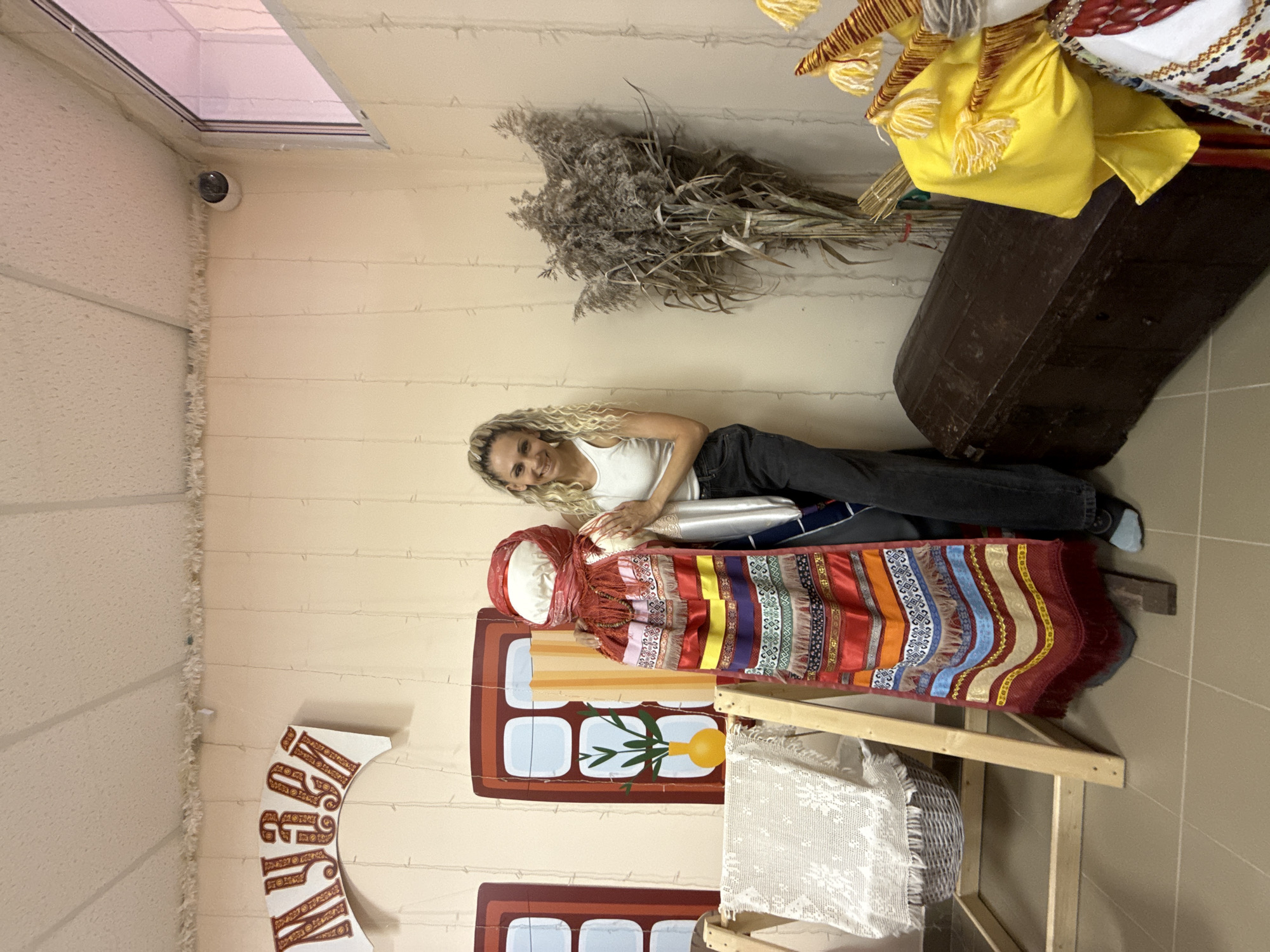 A woman with curly blonde hair, wearing a white tank top and black pants, lies horizontally on a wall decoration that imitates a lying doll. The decoration is colorful with various stripes and patterns, and is positioned on a wooden structure resembling a table or display stand. Behind the woman, on the wall, is a bundle of dried grass and a large, dark-colored object resembling a trunk. The wall itself is a light beige color with horizontal lines, and there's a window at the top left corner of the image. A CCTV camera is mounted near the window. The location appears to be a museum or exhibit in Шебунино, Russia, based on the exhibit style and the presence of Russian folk art and decorations.
