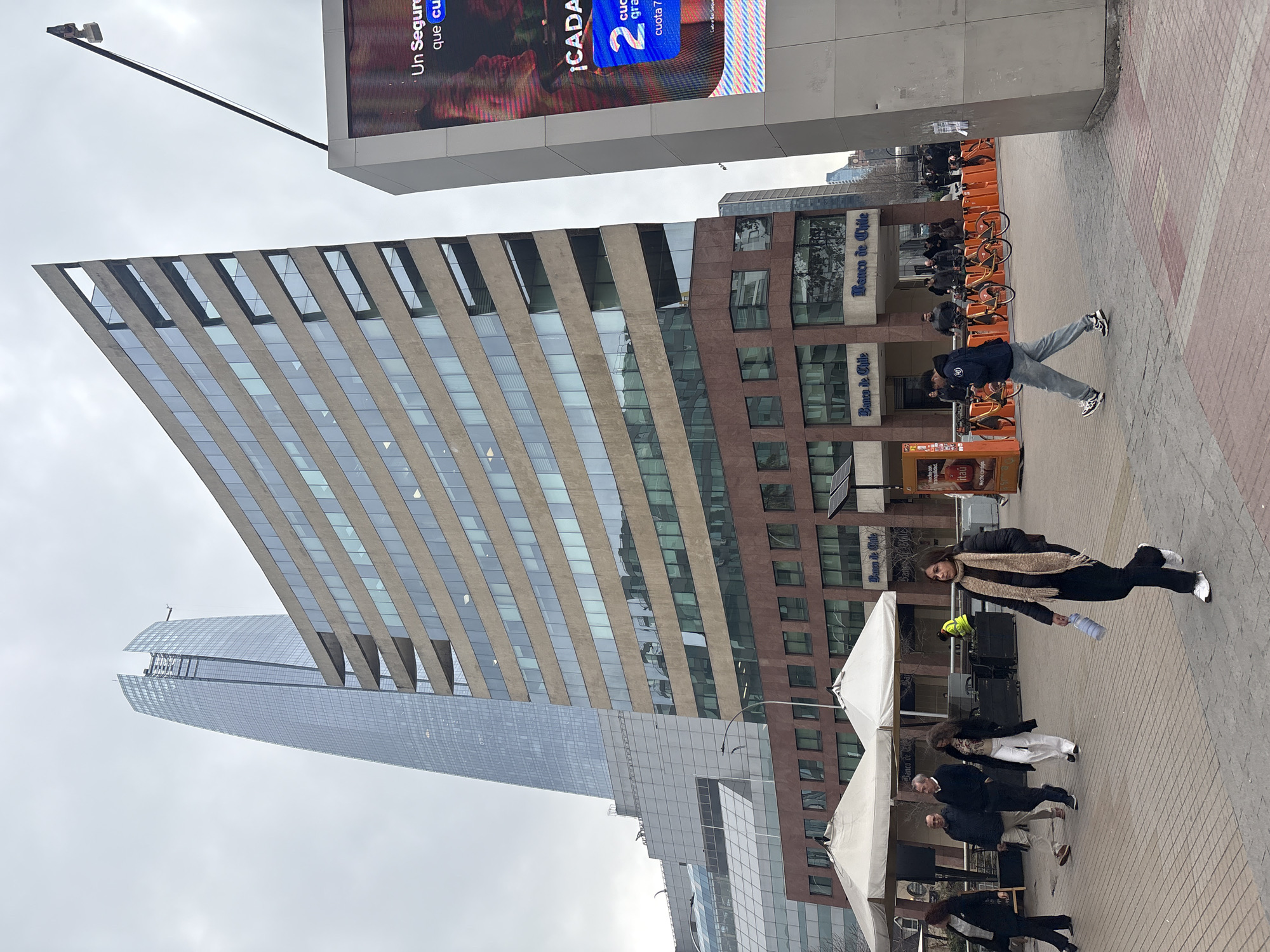The image depicts a street scene in Santiago, Chile, with the prominent Costanera Center skyscraper in the background. In the foreground, a modern, curved office building with a glass facade and beige stone accents dominates the view. To the right, a large digital billboard displays an advertisement with the text "Un Seguro que cuida". Several people are walking on a paved plaza in front of the buildings. Some are dressed casually, while others appear to be in business attire. In the middle ground, there are white tents, likely for outdoor vendors or events, and bicycle rental stations with orange bikes. The sky is overcast, suggesting a cloudy day.