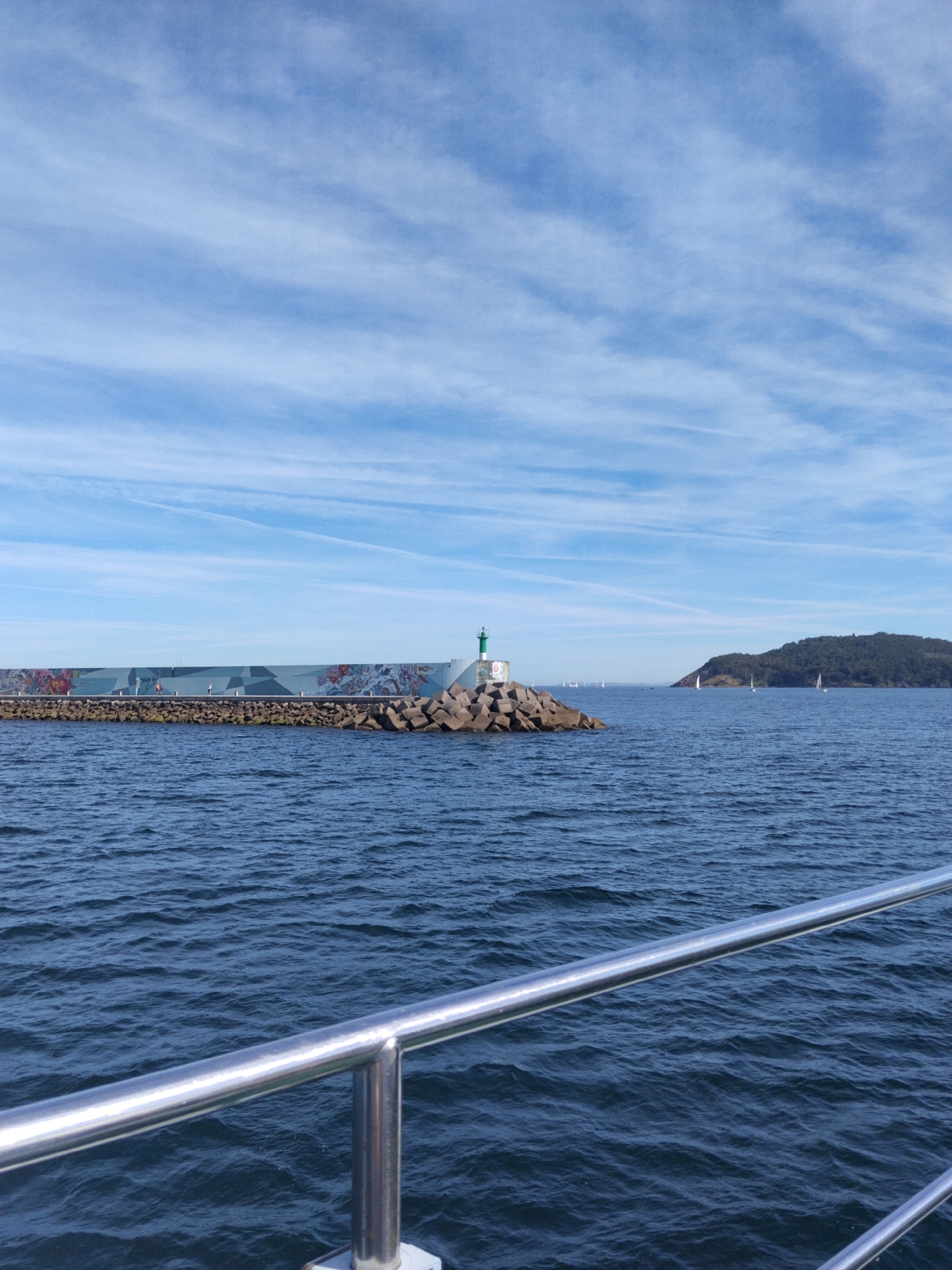 The image depicts a seascape with a breakwater adorned with colorful murals and a lighthouse at its end, located in the city of Baiona, Spain. The breakwater is constructed from large stones and concrete blocks. Several sailboats are visible in the distance, and a verdant, tree-covered island or hill rises from the sea on the right side of the frame. The sky is blue with wispy white clouds, suggesting fair weather. In the foreground, a metallic railing, likely part of a boat, spans the bottom of the image. The water is a deep blue with gentle waves.