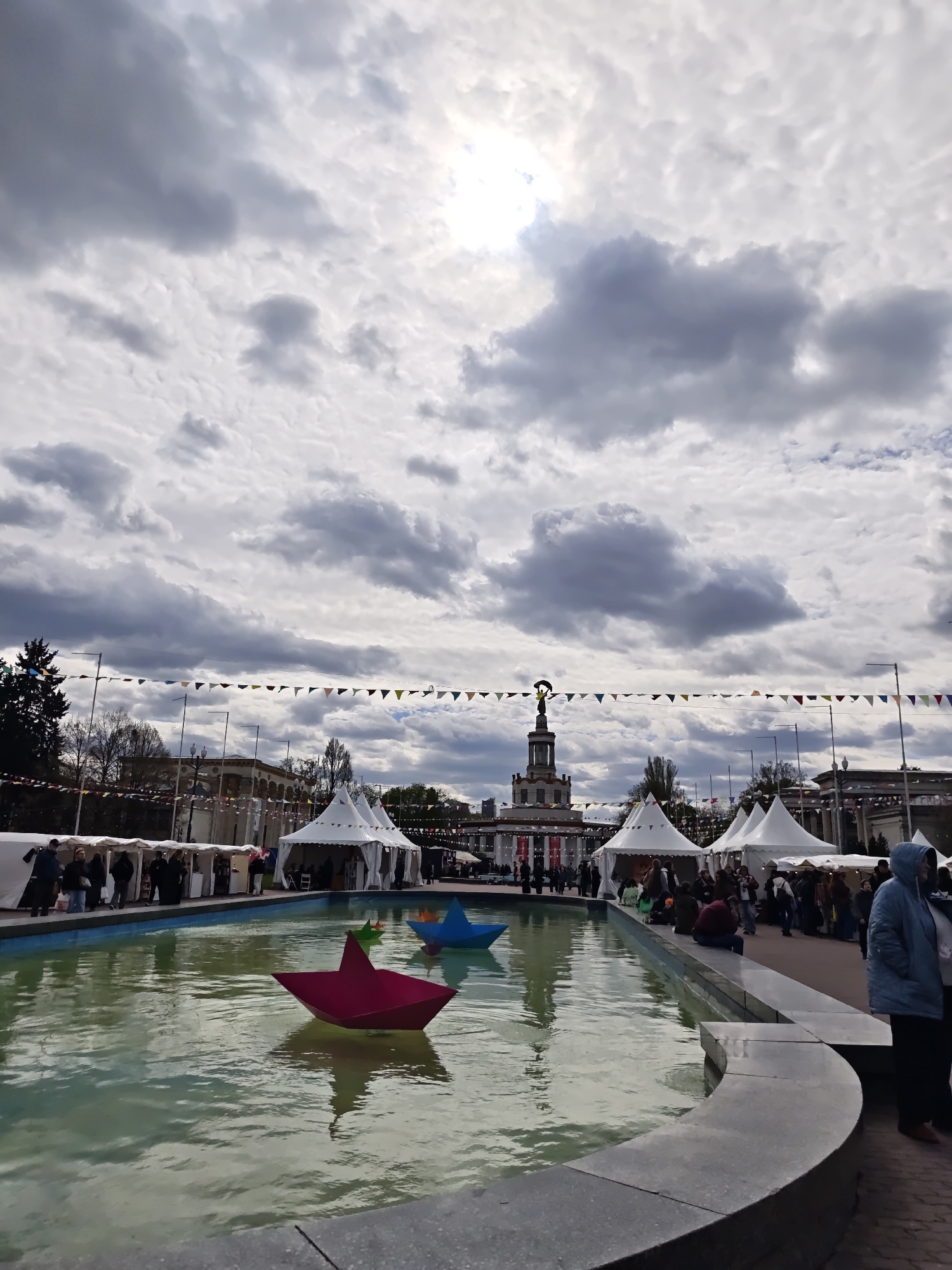 This media file captures an outdoor event at the Expocenter of Ukraine (VDNH) in Kyiv, characterized by a lively public gathering. The foreground features a large, rectangular water basin or fountain, reflecting the cloudy sky. Floating prominently in the water are several oversized, vibrant origami-style boats: a bright red one closest to the viewer, a blue one positioned behind it, and a smaller green one further back.
Surrounding the water feature, numerous white, conical tents or marquees are set up, suggesting market stalls, exhibition booths, or food vendors. A significant number of people are present, milling around the tents, congregating in groups, and some individuals are seated on the stone ledge bordering the water. One person in a light blue hooded jacket is visible on the far right.
In the midground, a grand architectural building with a distinctive tower topped by a statue serves as a prominent landmark, typical of the main pavilion at VDNH. Colorful triangular bunting is strung across the scene, connecting various poles and structures, enhancing the festive atmosphere. The sky is heavily clouded with a mix of dark and lighter grey clouds, but a bright patch in the upper center indicates sunlight breaking through. The overall impression is that of a bustling festival or fair taking place on a cloudy but bright day, likely in the late morning or afternoon. No visible text is discernible.