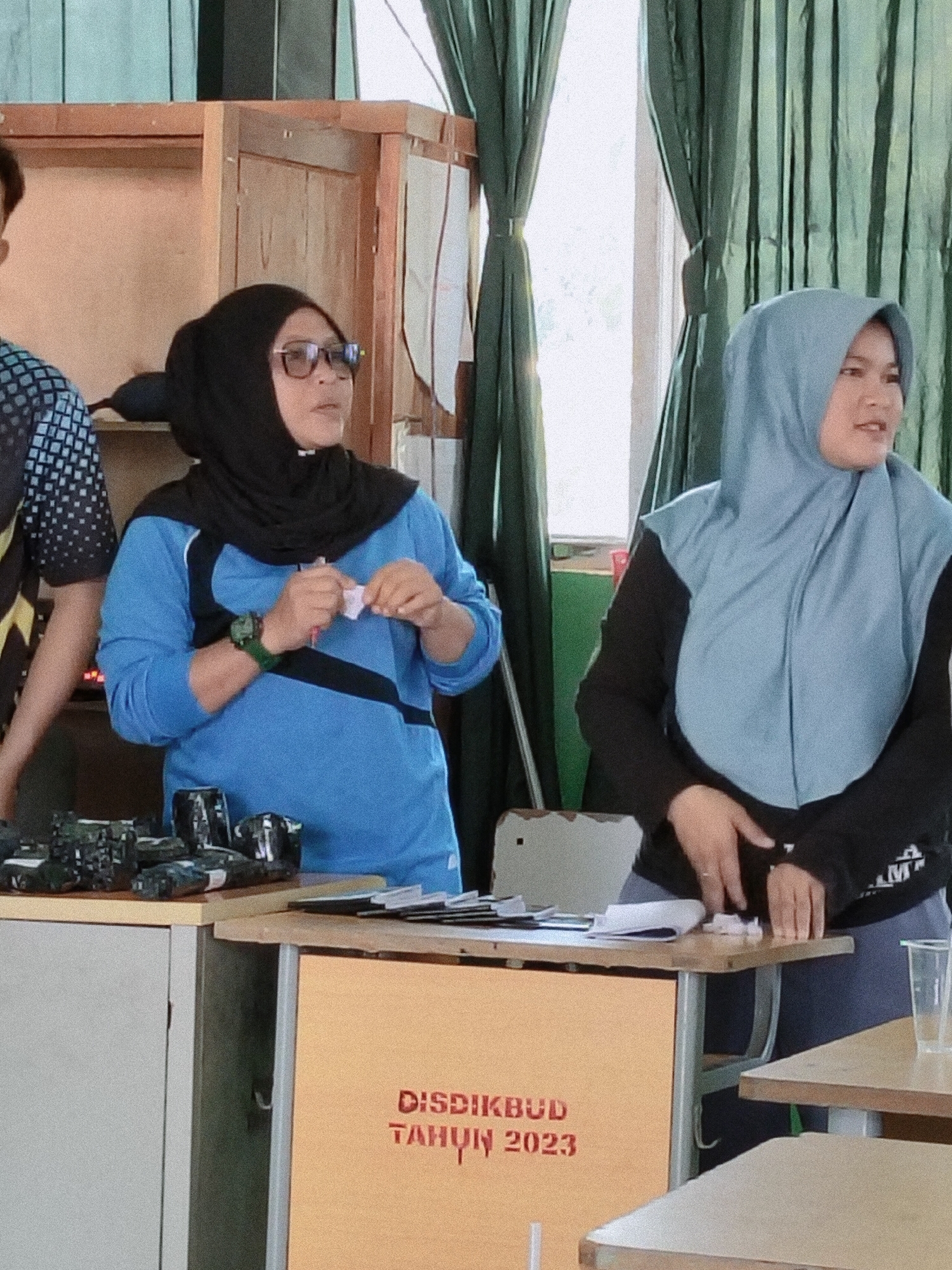 The image shows two women wearing hijabs in what appears to be an indoor setting with green curtains in the background. The woman on the left is wearing a blue long-sleeved shirt and a black hijab, and she is holding a small white object in her hands. She is also wearing glasses and a watch on her left wrist. The woman on the right is wearing a light blue hijab and a black long-sleeved shirt. Both women are looking to their right, suggesting they are observing something or someone outside the frame. In the foreground, there is a table with various items, including what look like small black objects wrapped in plastic, papers, and a clear plastic cup. A portion of a wooden cabinet is visible on the left. A sign on an orange panel in the lower part of the image reads "DISDIKBUD TAHUN 2023" in red lettering. The overall impression is a scene of an indoor event or meeting.