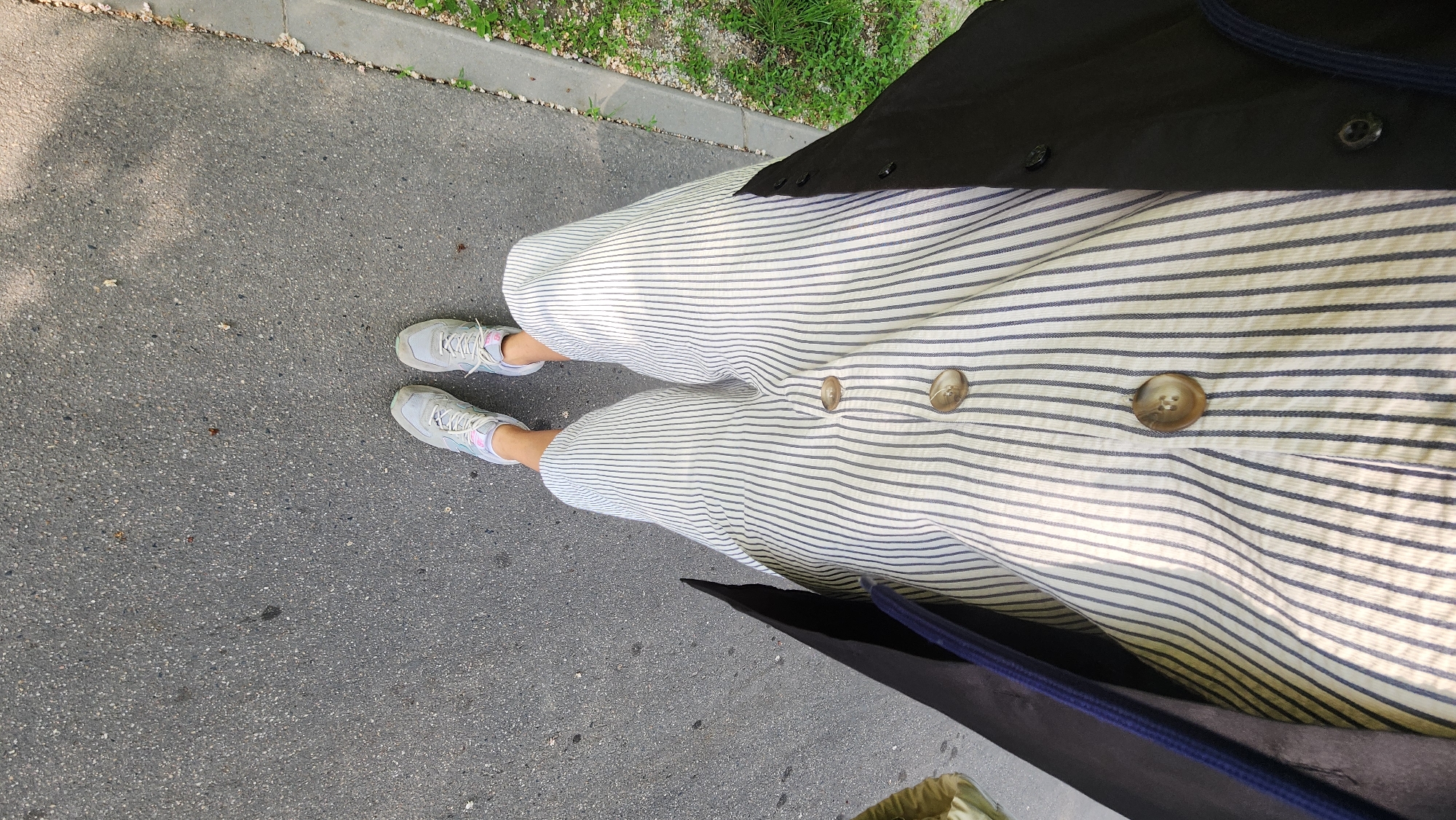 A first-person perspective, downward-facing full shot shows a person standing on a paved surface, likely a street or sidewalk. The person is wearing light gray athletic shoes and vertically striped, wide-leg pants with large brown buttons. A dark-colored coat or jacket is draped over the person's shoulders. The background includes a curb with grass growing beyond it. The setting appears to be urban, possibly a residential or commercial area.