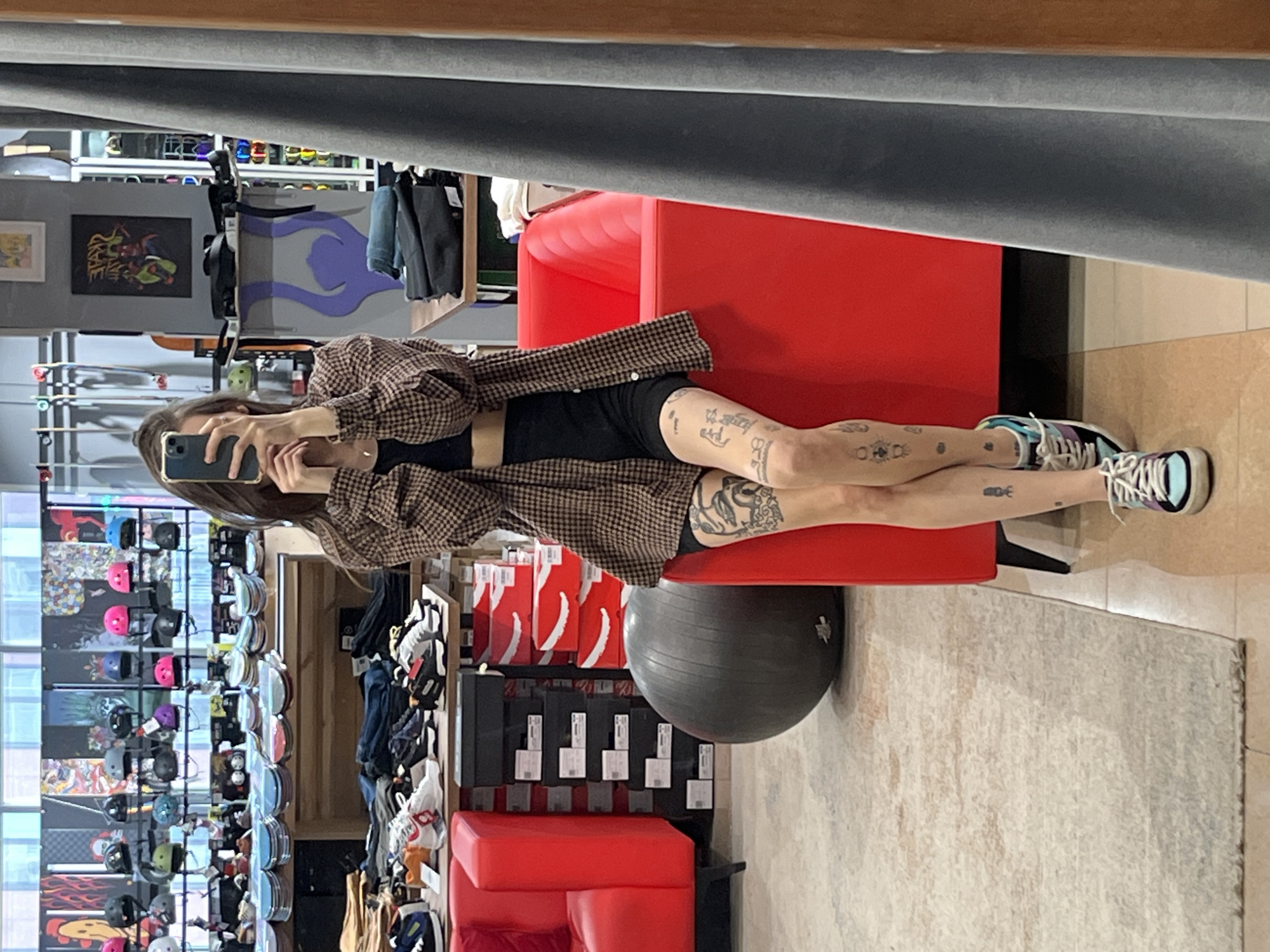 A woman is taking a mirror selfie while sitting in what appears to be a store. The store sells skateboards, shoes, and accessories. There are red benches, a gray exercise ball, and shelves of items. The location is a skate shop, possibly in Krasnoyarsk, Russia.