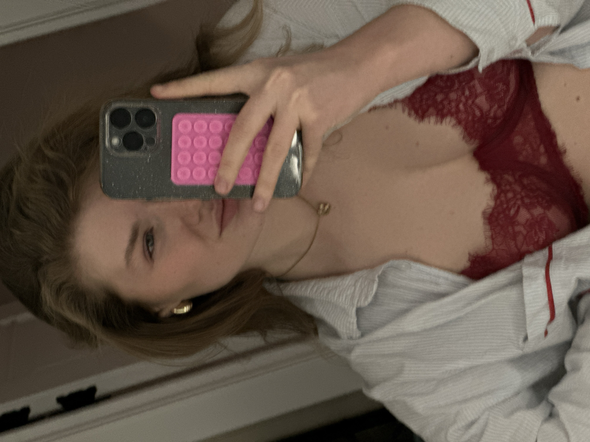 A woman with blonde hair takes a selfie in a mirror. She is wearing a red lace bra and a striped button-down shirt. The camera is partially obscuring her face. She is wearing a gold necklace and earrings. The location cannot be determined based on the image.