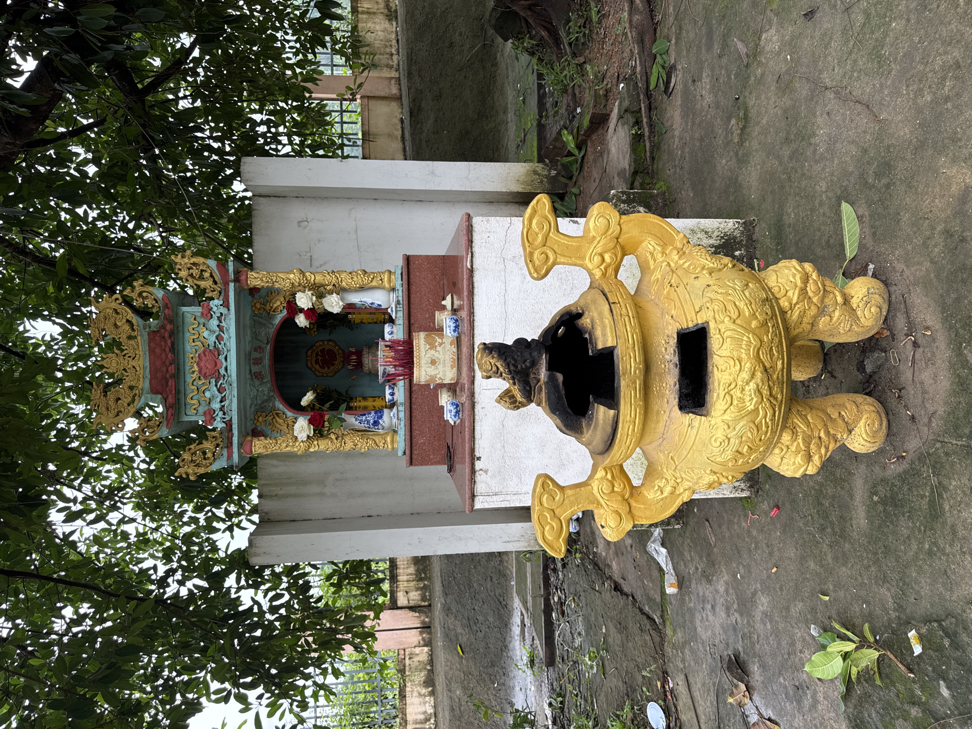 The image depicts an outdoor altar with a golden incense burner in the foreground and a multi-tiered shrine behind it. The shrine is adorned with colorful decorations, including red and gold motifs, and houses a circular emblem, red and white flowers in a vase, and incense sticks. The altar is situated on a concrete surface, which appears weathered and slightly overgrown with moss in some areas. Surrounding the altar are scattered leaves, debris, and a chain-link fence in the background. Overhead, lush green tree branches partially obscure the sky. The overall setting suggests a place of worship or remembrance in a somewhat neglected outdoor environment. The specific location in Lao Bảo Commune, Vietnam, is not visually identifiable from the image alone.