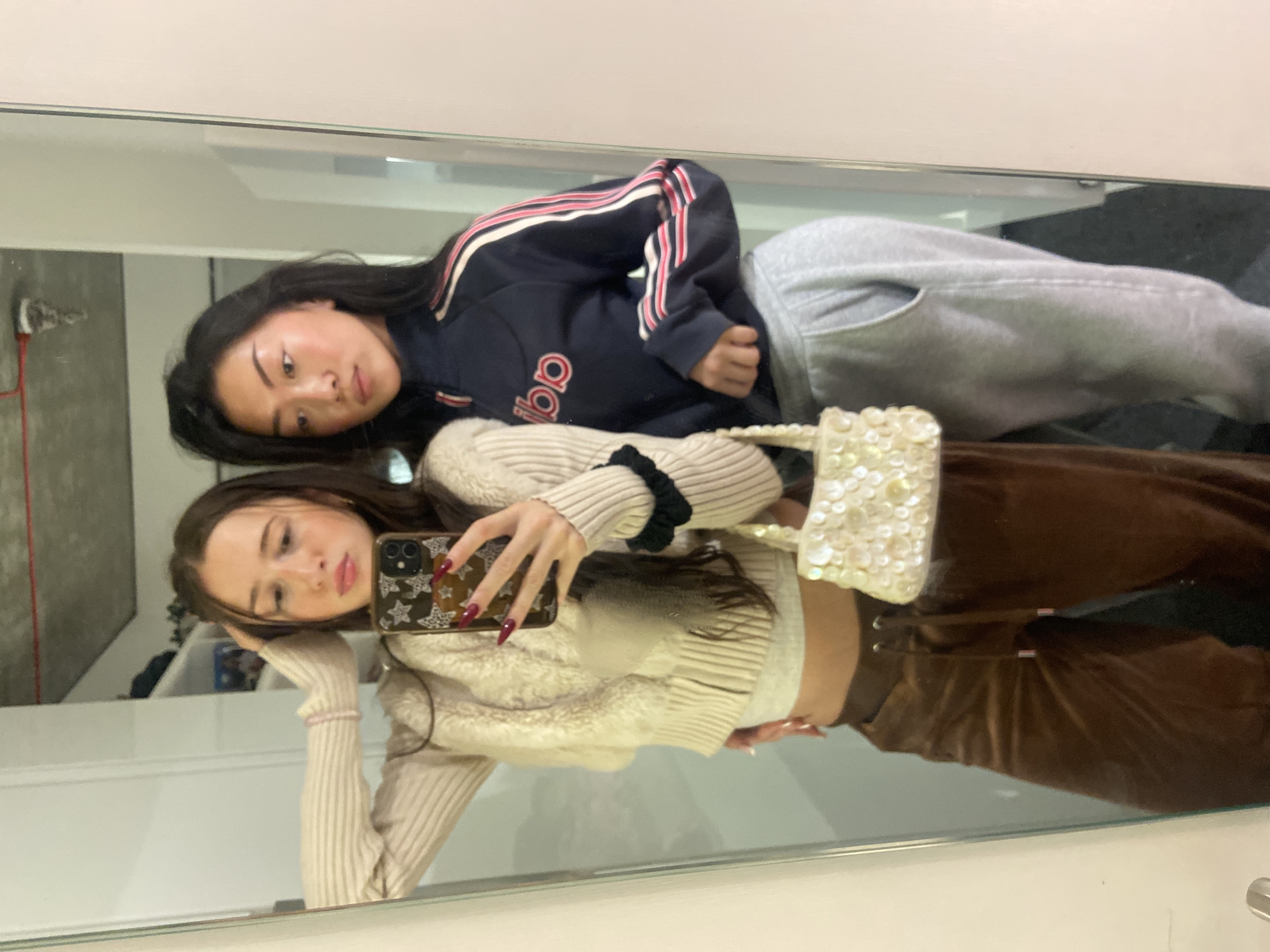 Two young women are posing for a selfie in a mirror. One woman, at the top of the frame, is wearing a navy blue Adidas sweatshirt with pink stripes and grey sweatpants. The other woman, in the foreground, is holding up a phone in a brown star-patterned case and wearing a beige sweater with brown pants and a small beaded purse. The background is a blur, but appears to be a room with white walls. Location is not discernible from the image.