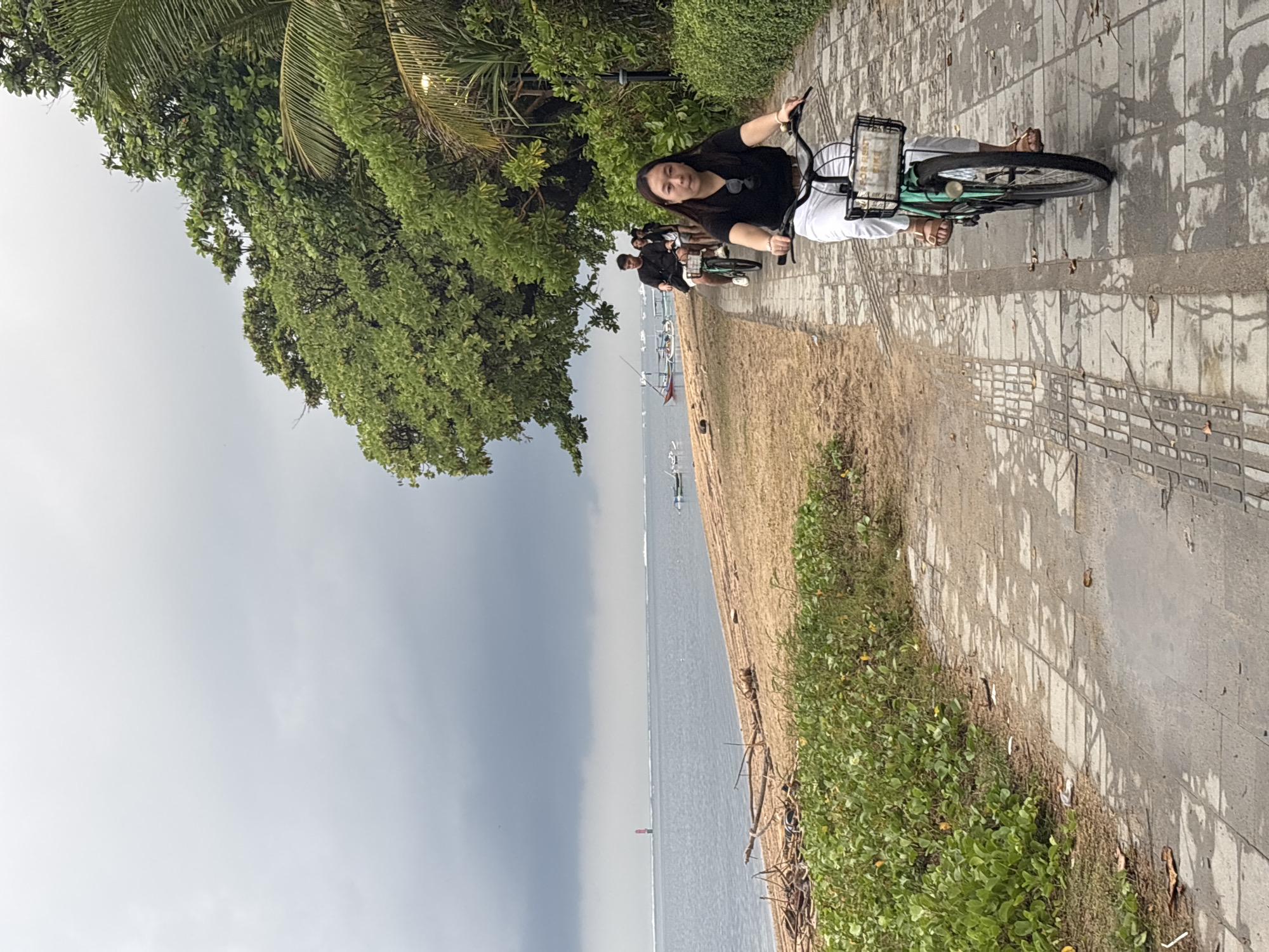 This image depicts a scene along the coast of Denpasar, Indonesia, on a cloudy day. In the foreground, a woman is riding a bicycle with a basket on the front, wearing a black t-shirt and white pants. She is on a paved pathway that runs parallel to a sandy beach. Behind her, another person is riding a bicycle, and further in the distance, a group of people are also on bicycles. The beach is lined with green vegetation and scattered driftwood. The ocean is visible, with a red buoy in the distance and several small boats. Lush green trees and foliage border the pathway on the right side of the image. The overall atmosphere is overcast, with dramatic clouds in the sky.