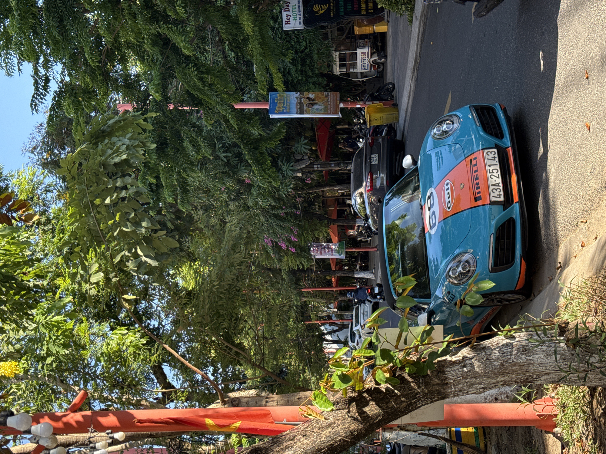 The image shows a street scene in Đà Nẵng, Vietnam, with a blue and orange Porsche 911 parked on the side of the road. The car has the number 68 and the Gulf logo on its hood. The license plate reads "43A-251.43".
The street is lined with trees, providing shade. A sign for the "Hey Day Hotel" is visible, along with a small food cart. Other parked vehicles and pedestrians are also present.
The lighting suggests it's daytime, with clear skies.