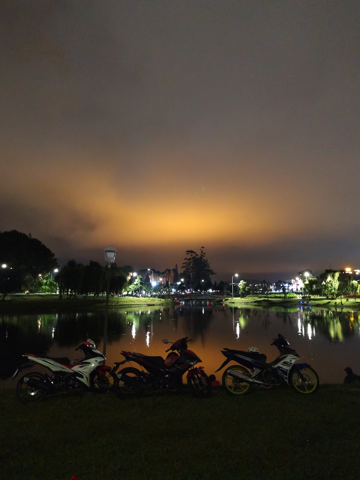 Three motorcycles are parked on the grass near a lake at night. The motorcycles are facing towards the lake. There is a street lamp behind the motorcycles. There are trees and buildings across the lake. The sky is cloudy and dark. The lights from the buildings are reflecting in the lake. There is a person sitting on the grass to the right. This is most likely in the city of Da Lat, Vietnam.