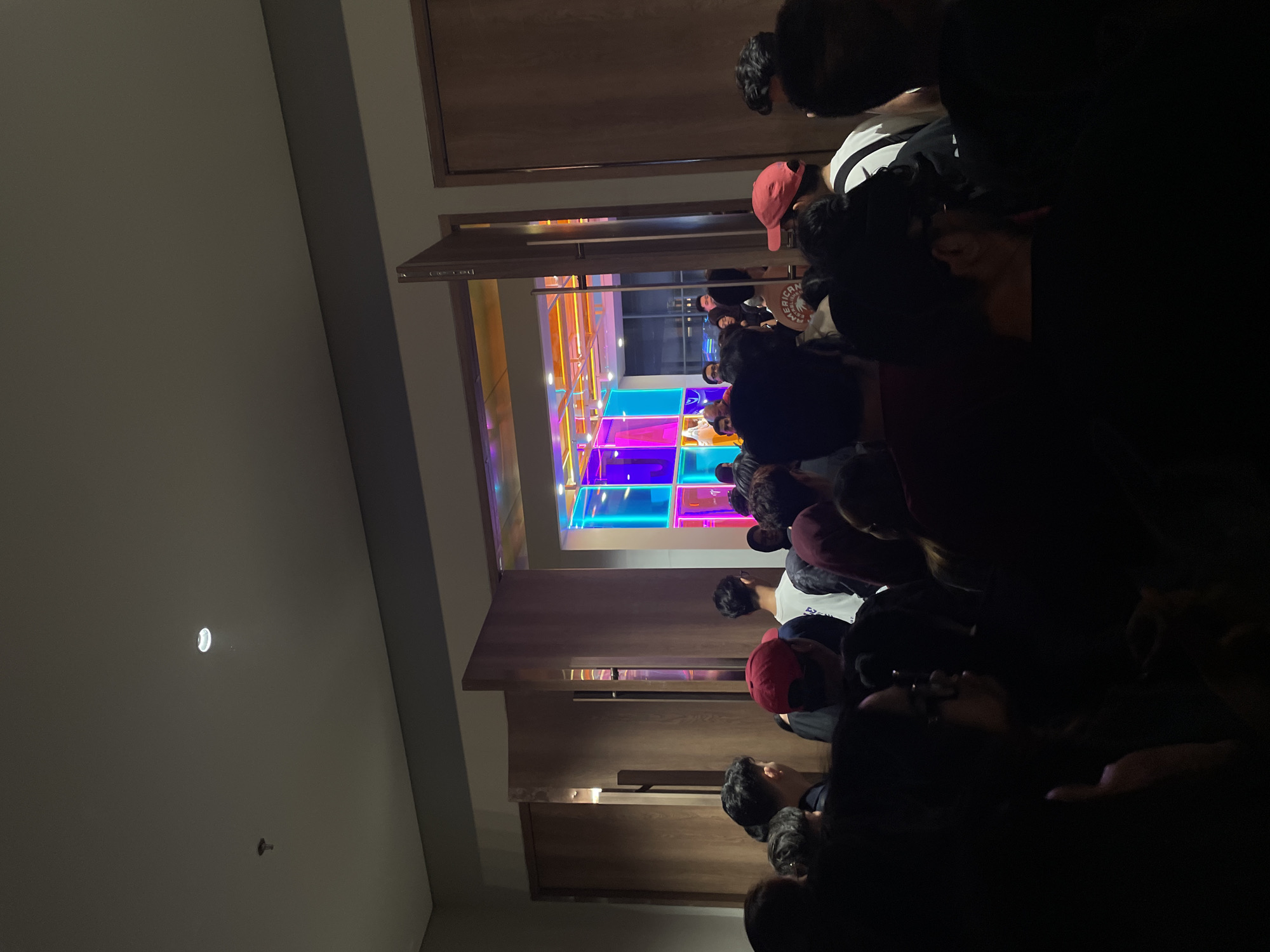 The image depicts a gathering of people in what appears to be an entertainment venue in Salembaran, Indonesia. The room features a colorful, illuminated floor composed of squares in shades of blue, pink, orange, and yellow. People are standing in a line or gathered in the room, but the view is partially obstructed by individuals in the foreground. The lighting is dim, with a strong contrast between the illuminated floor and the darker areas of the room and people. Wooden architectural elements, possibly door frames or wall panels, are visible around the opening of the space.