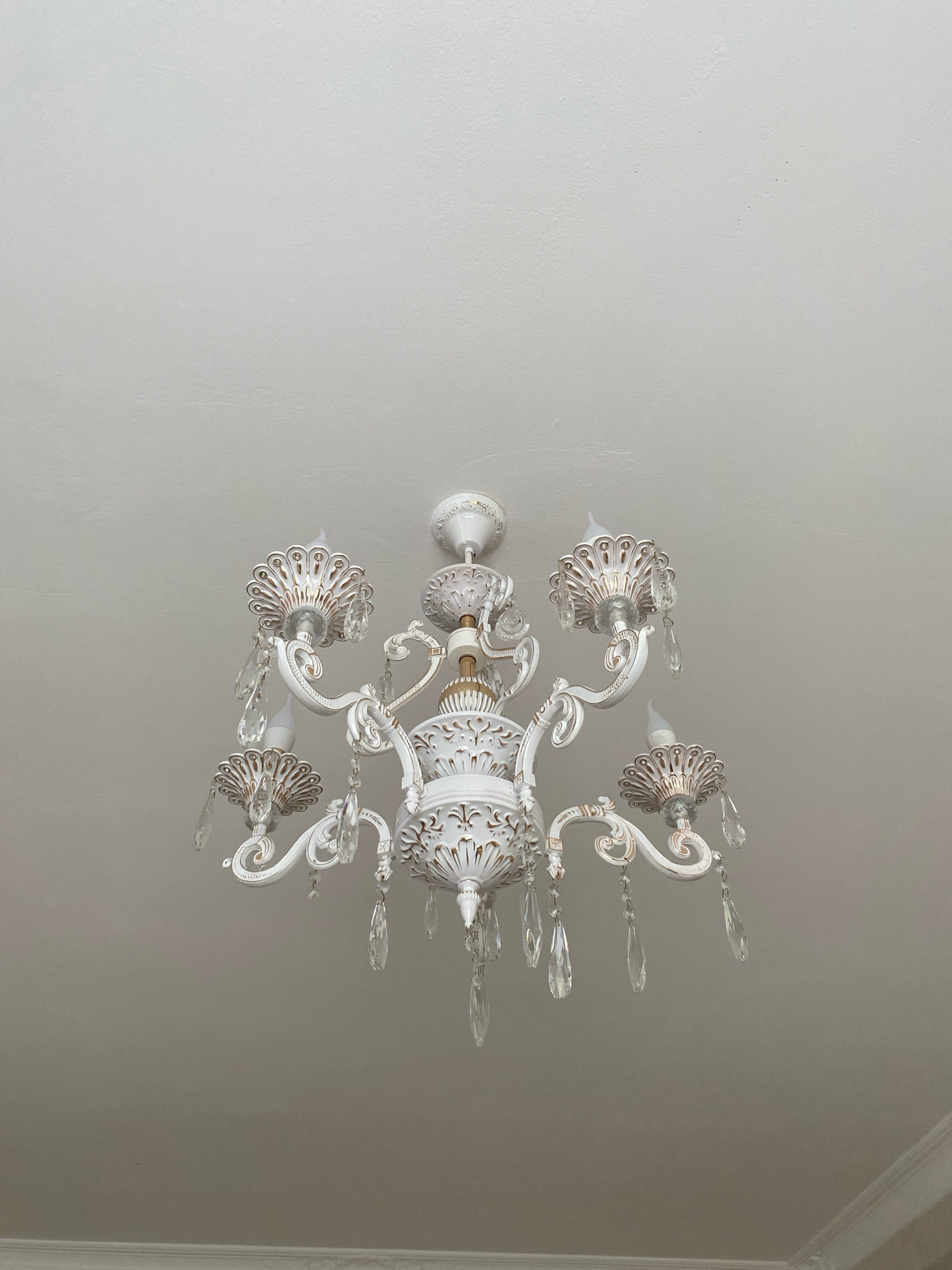 A white chandelier with gold accents hangs from a white ceiling. The chandelier has multiple arms, each adorned with crystal teardrop-shaped pendants and shell-like accents with gold detailing. The ceiling appears to be textured, and there is a decorative white trim along the bottom edge of the frame, suggesting the presence of a wall or upper structure. The lighting is neutral, illuminating the chandelier and the ceiling evenly. The image does not contain any people or specific environmental markers that would indicate a particular location.
