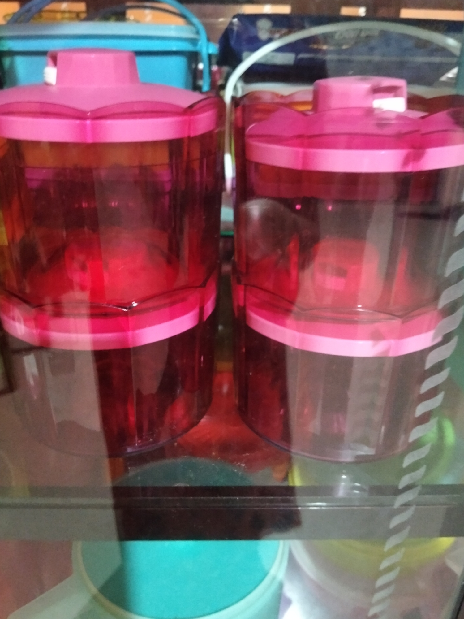 The image depicts two identical pink-lidded, translucent red plastic containers stacked on a reflective surface, likely a glass shelf. The containers have octagonal bases and are cylindrical in the middle section. They appear to be storage containers. In the background, a bright blue plastic container with a handle is visible, along with other out-of-focus objects. The lighting is dim, and the overall scene has a somewhat cluttered appearance. The image does not provide any specific details about the city of Payagaboh, Indonesia.