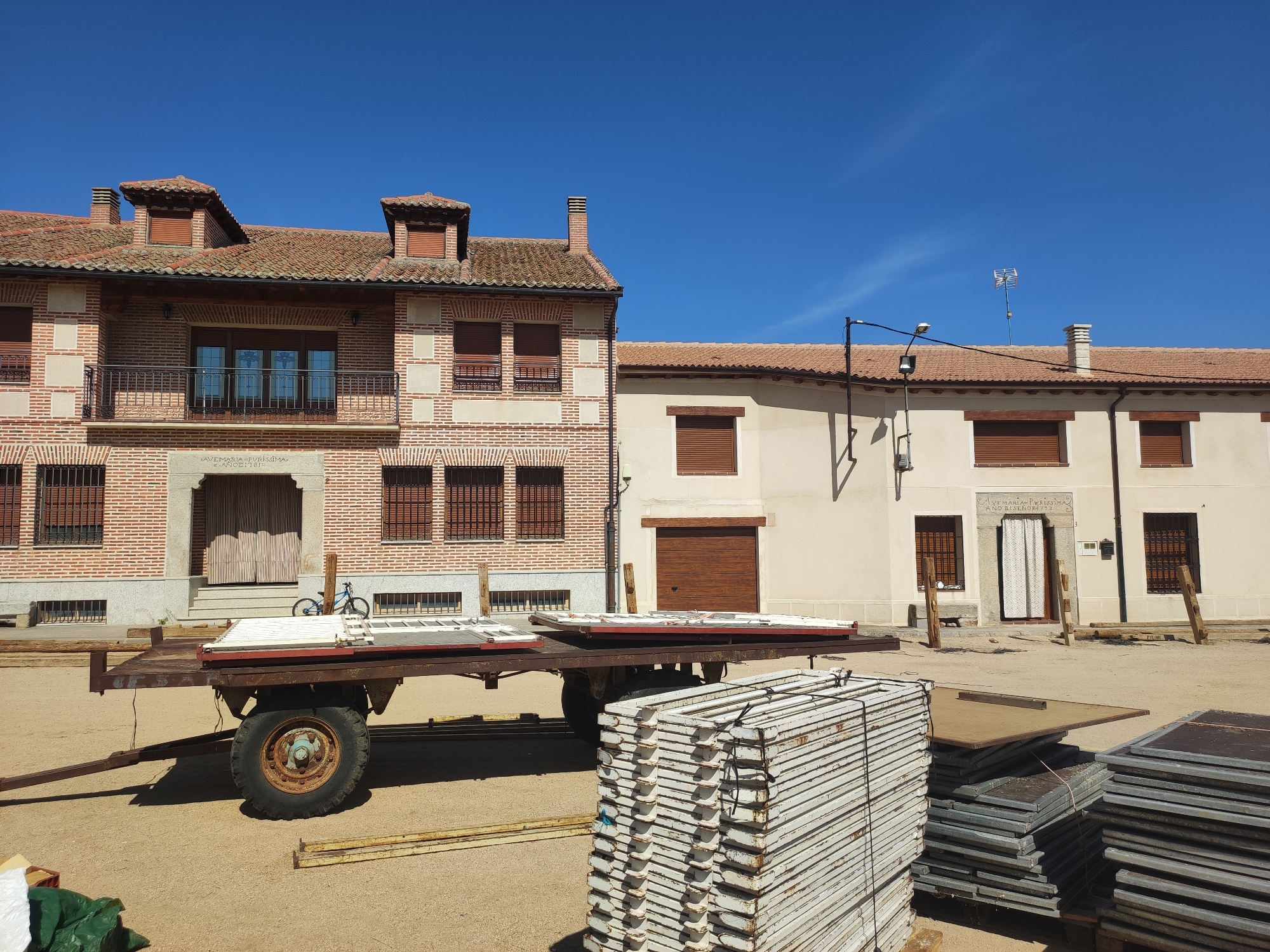 The image shows a daytime outdoor scene in Sangarcía, Spain. In the background, there are two connected buildings with terracotta tiled roofs. The building on the left is made of red brick with lighter stone accents and features multiple windows with brown shutters, a balcony with a wrought-iron railing, and dormer windows on the roof. The building on the right is lighter in color, with a similar tiled roof, brown shutters on its windows, and a large wooden garage door. A blue sky with some faint wisps of clouds is visible above.
In the foreground, a large, rusty flatbed trailer is parked on a sandy or gravelly surface. The trailer has one visible large wheel with a light blue hub. It is loaded with several long, flat white objects, possibly scaffolding planks or large panels. To the right of the trailer, a large stack of flat, metallic objects, possibly more panels or metal sheeting, is neatly piled. Next to these, another stack of similar items is also visible. There are also some loose wooden beams or planks scattered on the ground. A blue bicycle is leaning against the side of the brick building, near the trailer. The overall impression is a construction or renovation site.
