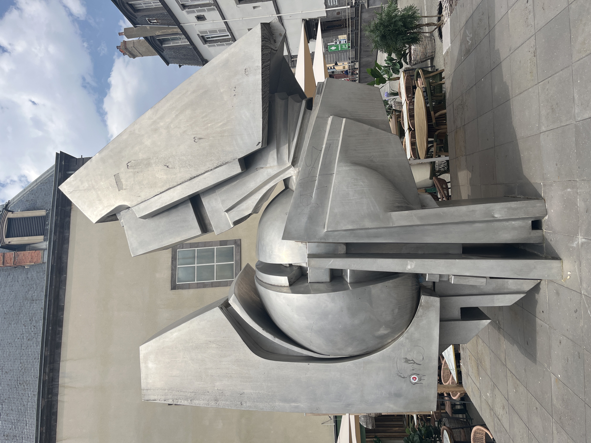 This is a photograph of a large, abstract metal sculpture located in a plaza in front of buildings in Clermont-Ferrand, France. The sculpture is composed of interlocking geometric shapes and a central sphere, all rendered in brushed metal. The plaza pavement is made of gray square tiles. In the background, there are buildings with visible architectural details such as windows and chimneys. Outdoor seating areas with tables and chairs are visible, some with umbrellas. A few potted plants are also present. The sky is partly cloudy. The sculpture is the primary subject of the image, dominating the foreground. No people are clearly identifiable in the immediate vicinity of the sculpture.