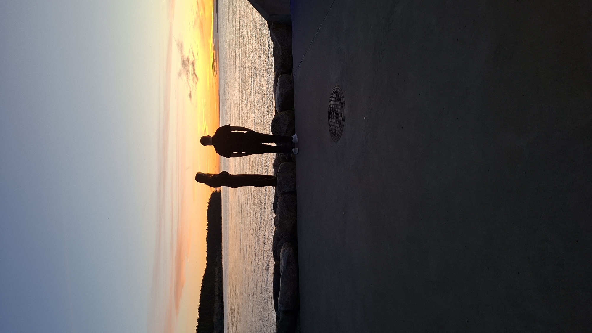 Two individuals, a woman and a man, are silhouetted against a vibrant sunset over the Baltic Sea. The woman, on the left, stands with her back to the viewer, her long hair falling behind her. The man, to her right, faces the water with his hands in his pockets. They are positioned on a concrete walkway that is lined with large, rough-hewn stones. In the foreground, the concrete walkway extends downward and is mostly in shadow, with a visible manhole cover. The background features a dark, tree-lined shoreline on the left and the expansive, calm water reflecting the colors of the sunset – oranges, pinks, and blues – in the sky. The scene appears to be a waterfront promenade. Without further information, the precise location in Tallinn cannot be determined.