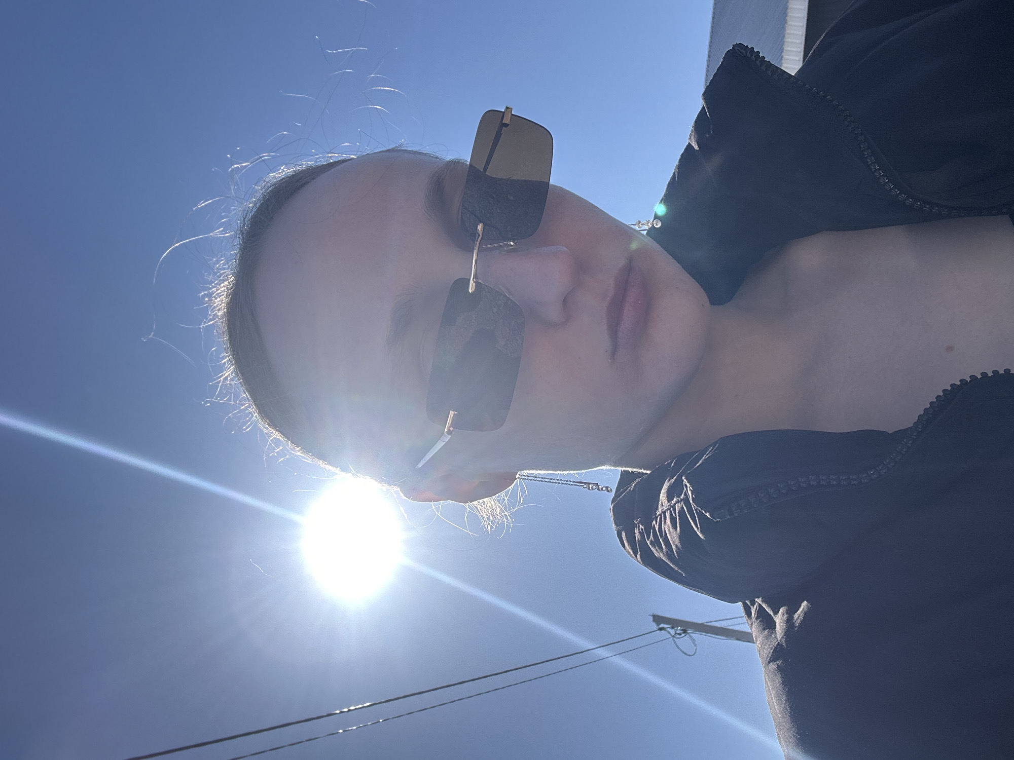 The image shows a person's face wearing sunglasses under a bright sun and blue sky. The person is wearing a dark-colored jacket. The location is not identifiable as Подшивалово, Russia from the given image. Utility poles and wires are visible in the background. The photograph appears to have been taken from a low angle, looking upwards toward the sky and sun.