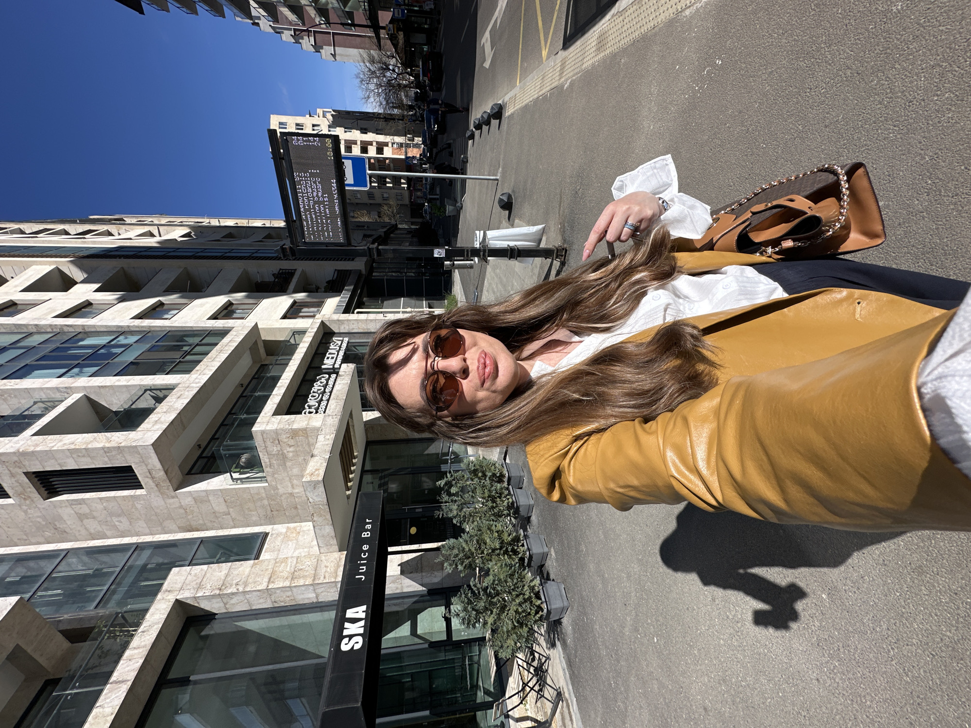The image shows a woman in sunglasses and a mustard-colored leather jacket, posing for a selfie on a city street. A tan leather shoulder bag with a chain strap is slung across her body. In the background, a modern building with the sign "SKA Juice Bar" is visible. A vertical LED display board displaying text is next to the building. The street appears to be in Tbilisi, Georgia.