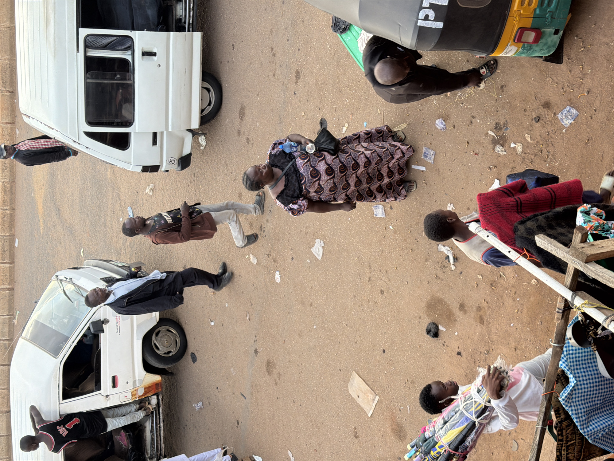 The image depicts an outdoor scene in what appears to be a marketplace or roadside in Bukuru, Nigeria. The perspective is from above, looking down on the subjects.
Several people are visible, some standing, some seemingly in transit. In the foreground, a person wearing a white shirt and black pants is standing next to a white van. Another person in a black tank top with the number "1" on it is leaning into the back of the same van, which has "POWER" and "SPORT" written on its side. A man in a blue shirt and black suit jacket, along with a person in grey pants and a red and black checkered jacket, are near another white van. A woman in a patterned dress with a black purse and a water bottle is also present, along with a person in a black outfit. In the background, a man in a green shirt is partially visible near a yellow and green auto-rickshaw. Another individual is seen sitting.
The setting is a dusty, paved area with scattered debris, including crumpled paper and other refuse, suggesting a busy public space. The lighting suggests it is daytime, with no clear indicators of specific weather conditions.
One person is holding a bundle of what appear to be umbrellas, suggesting they might be a vendor. The overall impression is of a bustling everyday scene, likely a commercial hub. The faces of the individuals are not clearly visible or expressive enough to ascertain specific emotions.
The text visible on the van reads "POWER" and "SPORT". The auto-rickshaw has "P1" and a partial logo visible. There is also a circular logo with "CH" on the rear of the first white van.