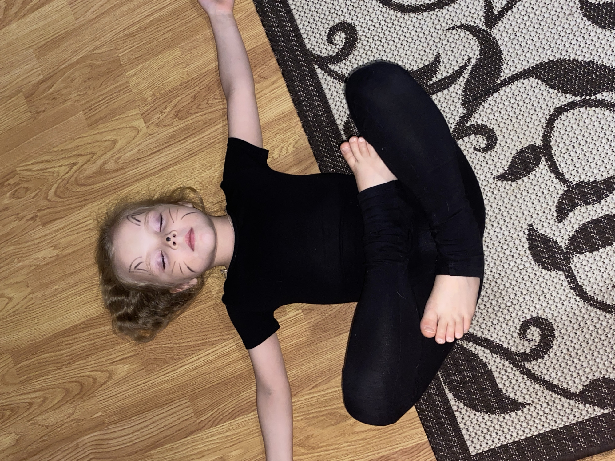 A young child is depicted lying on their back indoors, partially on a light brown wooden laminate floor and partially on a rug. The child has light brown, wavy hair and is wearing a black short-sleeved top and black leggings. Their face is painted with black whiskers on both cheeks and a small black nose, resembling a cat. Their eyes are closed, and their expression appears relaxed or playful.
The child's left arm is stretched upwards and outwards, while their right arm is bent with the hand near their chest. Their legs are bent in a flexible pose, with the left foot resting on the inner thigh of the right leg. The floor on the left side of the image features distinct wood grain patterns. On the right side, a rug with a light base color and a dark brown, ornate swirling pattern is visible. The lighting suggests an indoor setting, likely artificial. There is no visible text in the image. The scene conveys a moment of playful posing or rest. While not visible in the image, the context provided states the location is Votkinsk, Russia.