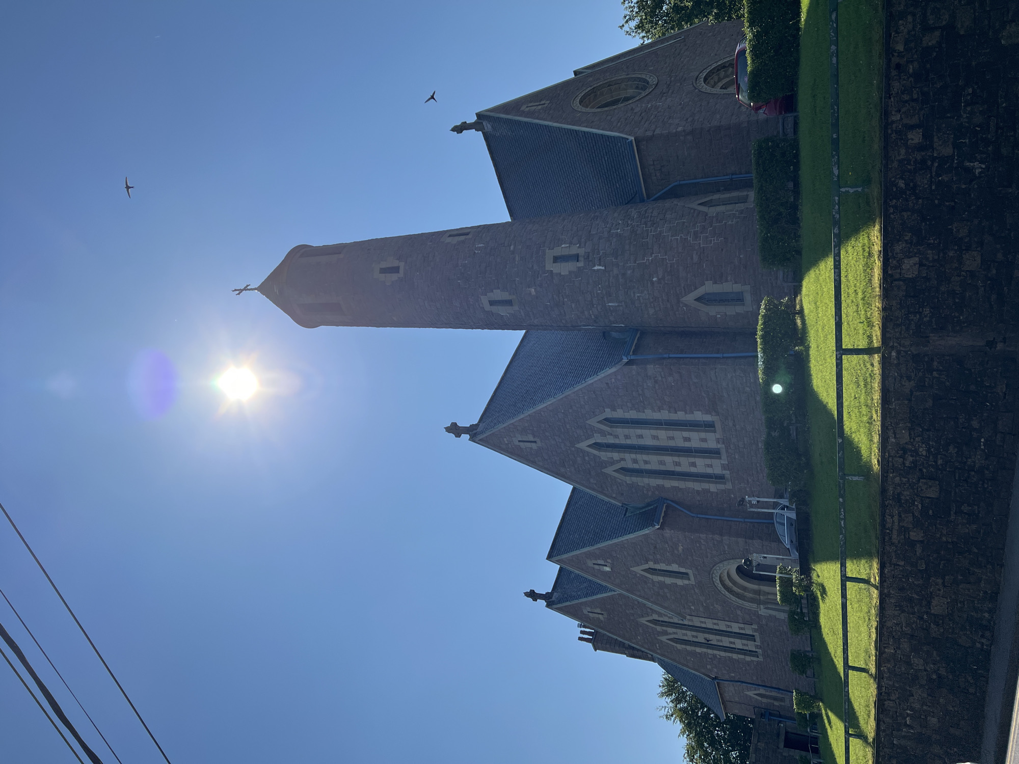 This image shows a stone church with a tall, round tower, bathed in bright sunlight under a clear blue sky. The church has a steep roof with multiple gables, each topped with a cross. The tower also has a cross at its apex. The sun is shining intensely, creating lens flare and casting long shadows across the neatly manicured lawn in front of the church.
There are two cars visible, one silver sedan partially obscured by bushes near the entrance and a red hatchback parked further back. A low stone wall runs along the bottom of the frame, separating the church grounds from a street or pathway. Two birds are visible flying in the sky. The scene appears to be a sunny day in a peaceful setting.
This church is likely the Church of Ireland parish church in Donegal Town, Ireland, officially known as Drumholm Parish Church. It is located on the west side of the town, overlooking Donegal Bay. The architecture is typical of Gothic Revival style, and the stone construction suggests a solid, long-standing building. The presence of the cars and well-maintained grounds indicates it is an active place of worship.