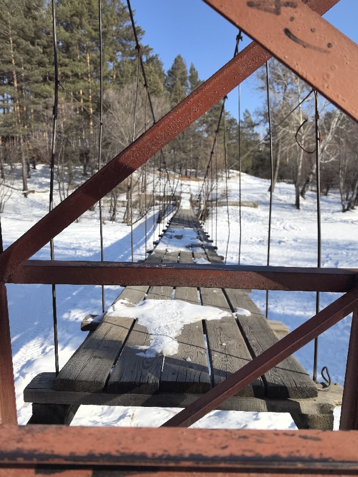 The image depicts a static scene of a suspension bridge during winter in Atamanovka, Russia. The viewpoint is from the immediate foreground, looking down the length of the bridge. Two rusty, dark red metal structural beams of the bridge, one horizontal and one diagonal, frame the lower and upper-right portions of the frame. A faint, hand-drawn smiley face is visible on the upper-right diagonal beam.
The suspension bridge features a wooden plank walkway, appearing weathered, with multiple parallel planks running longitudinally. Patches of white snow and ice are present on the wooden planks, most notably a distinct irregular patch in the lower-center of the bridge deck. Numerous dark metal support cables extend vertically and diagonally from the bridge's side rails, visible along its entire length.
The ground on both sides beneath and beyond the bridge is covered in a uniform layer of white snow. To the left, a dense stand of tall evergreen trees, likely pines, dominates the landscape. To the right, a sparser collection of bare deciduous trees with visible white bark is present. A bright, clear blue sky is visible above the tree line. The scene is illuminated by direct sunlight, casting distinct shadows from the bridge structure and trees onto the snow-covered ground. No individuals or active subjects are present in the frame.