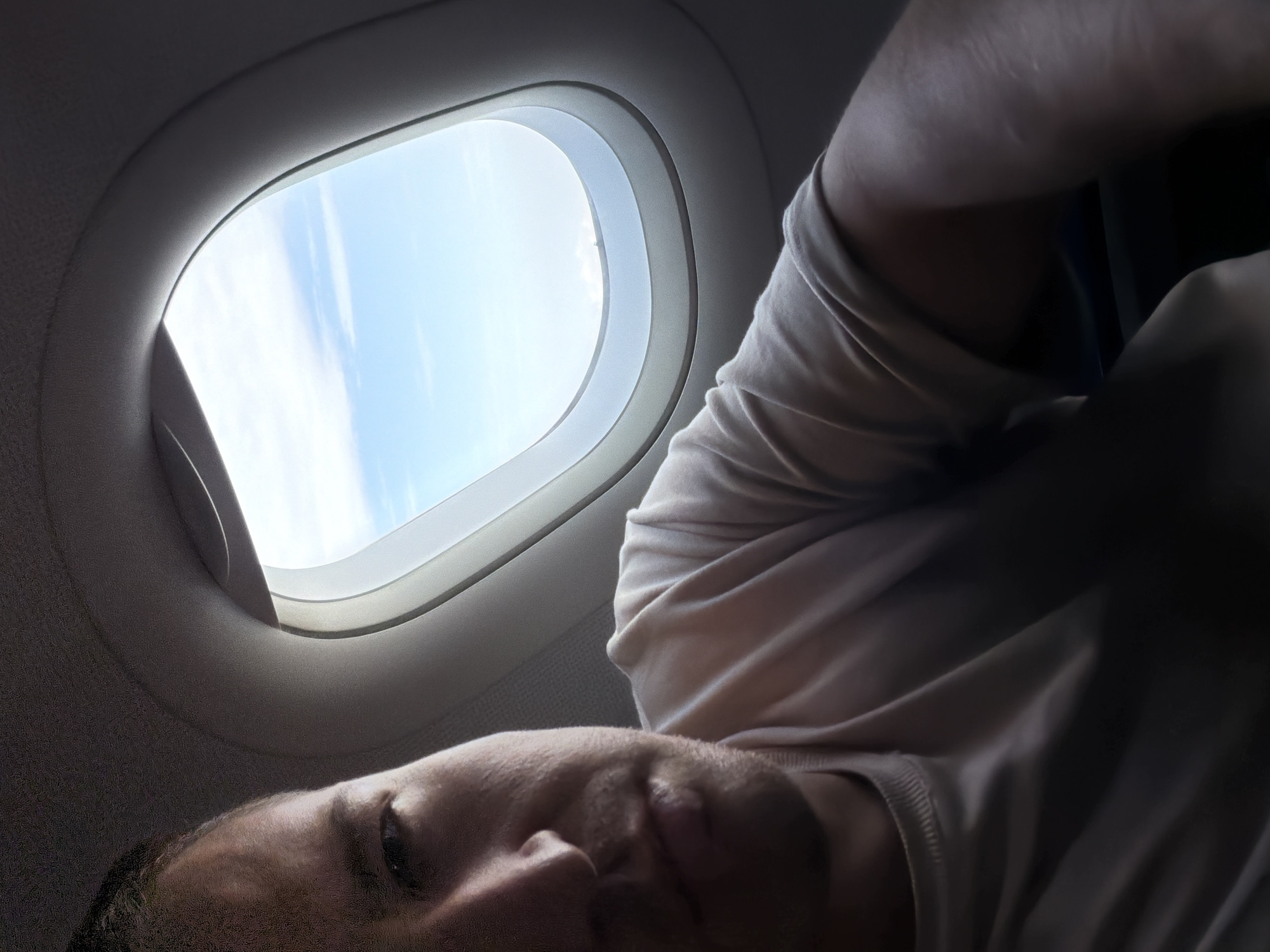A man is looking into an airplane window. The window is oval-shaped with a white frame. Outside the window, there is a clear blue sky with some wispy clouds. The man is wearing a light-colored t-shirt. The image is taken from a low angle, showing the man's face and upper body. There is no identifiable indication of location in the city of Mohan, India.