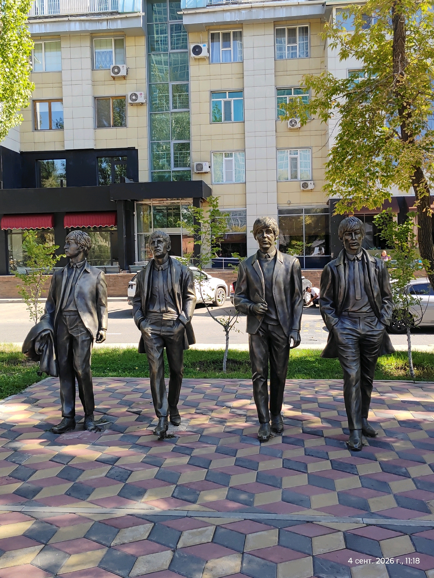 The image depicts a full shot of a street scene in Astana, Kazakhstan. In the foreground, four bronze statues of The Beatles are arranged in a line, facing the viewer. They are positioned on a decorative paved walkway with a geometric, illusionistic pattern of diamond-shaped tiles in shades of red, yellow, and grey. The statues are life-sized and depict the band members in their characteristic suits and hairstyles.
Behind the statues, a modern, multi-story building with a light-colored facade and numerous windows dominates the background. The building features air conditioning units on its exterior. A paved street runs parallel to the sidewalk, with a few cars parked on the far side. Green grass and small trees line the area between the statues and the street, and larger trees with green and yellowing leaves cast shadows on the scene. The sky is visible above the building, indicating an outdoor setting during the daytime. A timestamp in the bottom right corner reads "4 сент. 2026 г., 11:18," which translates to "September 4, 2026, 11:18." This suggests the photo was taken on this date and time.
