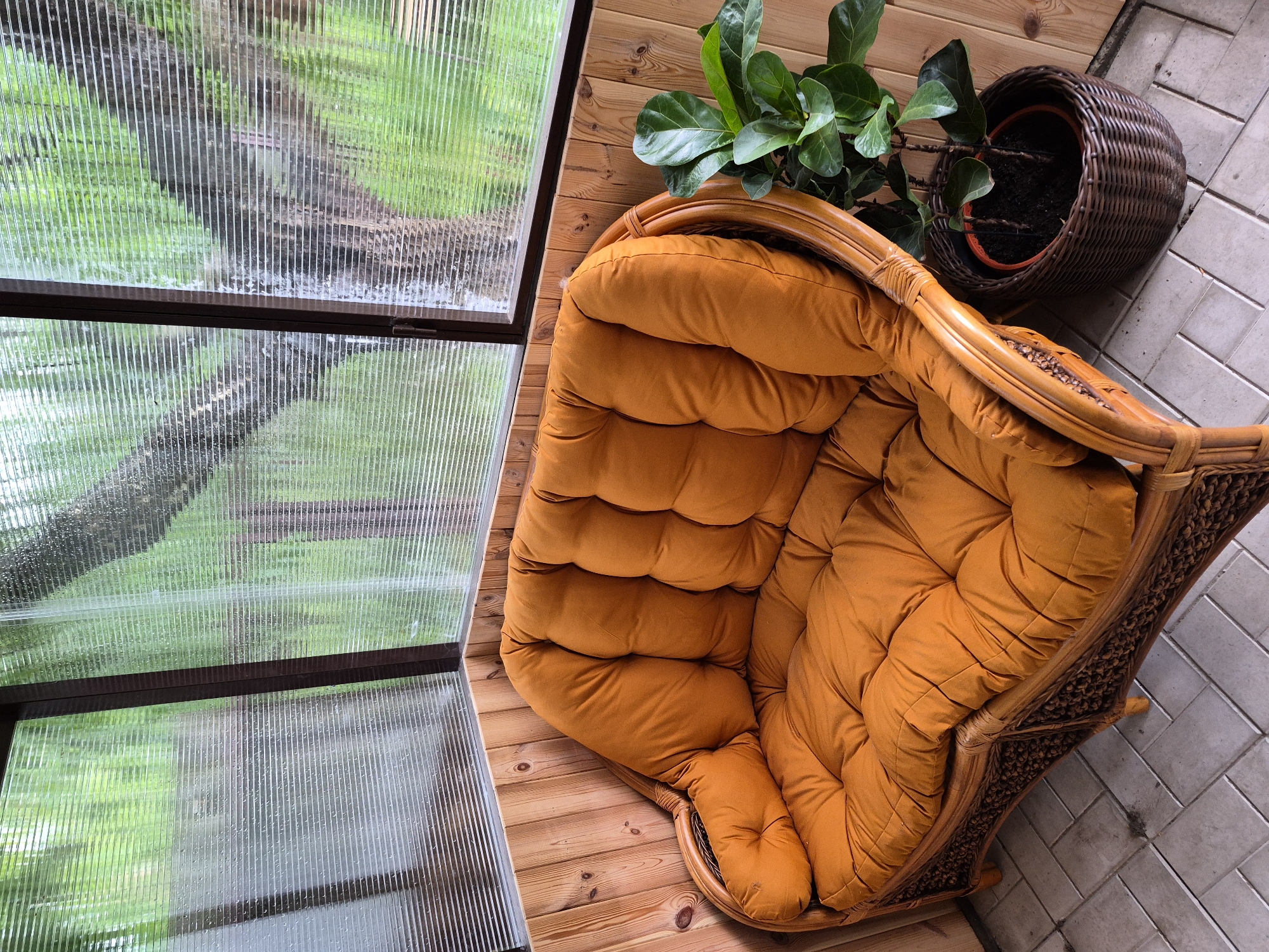The image depicts an enclosed corner of a structure, possibly a veranda or sunroom, located in Hlevakha, Ukraine. The structure features wooden paneling on the lower walls and corrugated, translucent polycarbonate or plastic sheeting for the upper walls and windows. The exterior surface of the window panels shows numerous small water droplets, indicating recent precipitation or condensation. Through the ribbed windows, a blurred view of dense green foliage and tree trunks is discernible. The floor is covered with light grey square tiles arranged in a herringbone or basketweave pattern.
A two-seater sofa occupies the corner, featuring a woven rattan or wicker frame in a light brown hue. The sofa is fitted with thick, tufted cushions of a rust-orange or burnt orange color on both the seat and backrest. To the right of the sofa, a plant is situated on the floor. The plant, possibly a Ficus lyrata given its large, dark green leaves, is contained within a terracotta-colored pot, which is then placed inside a dark brown woven basket. There are no individuals or discernible actions present in the scene.