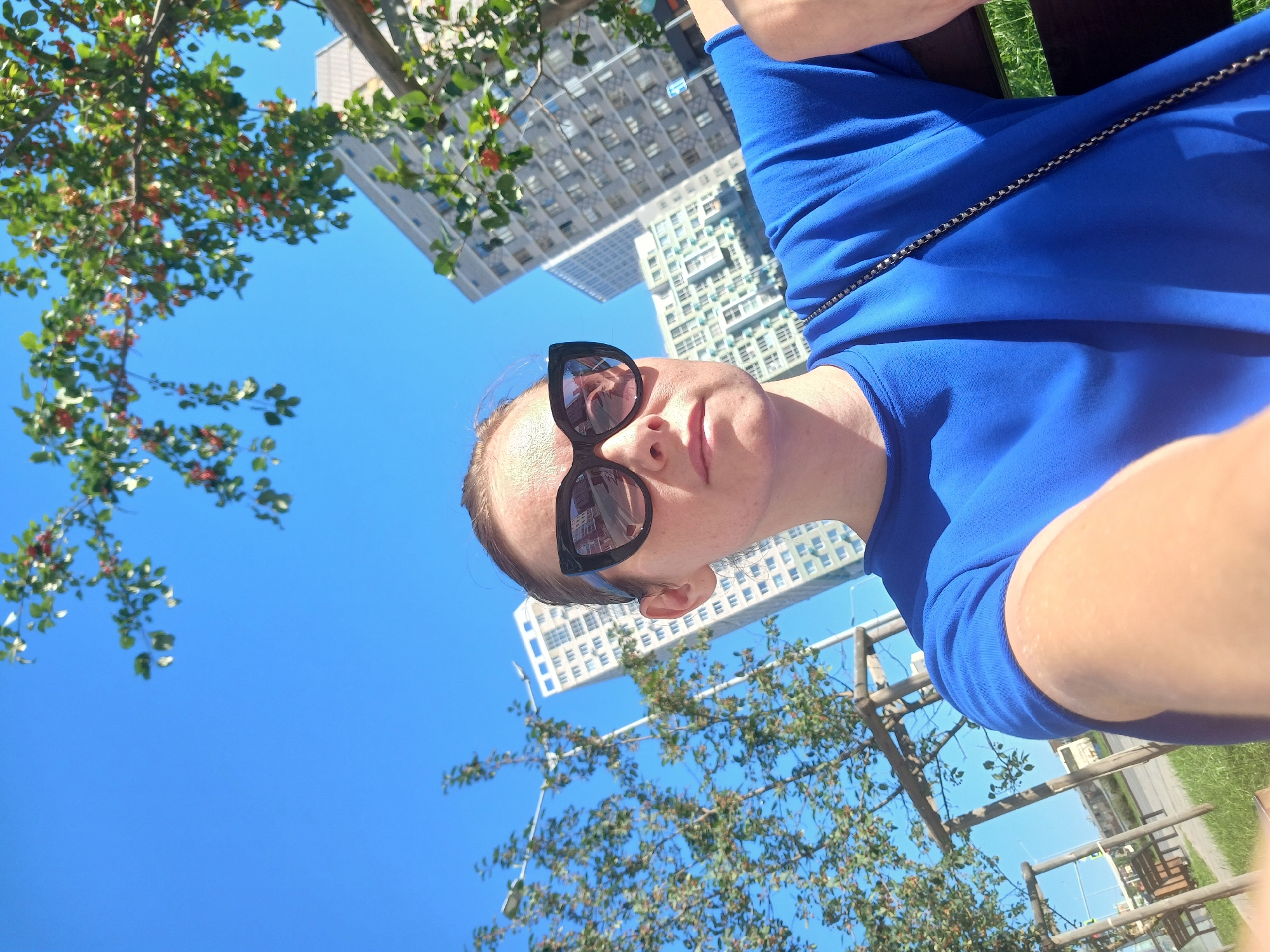 A woman in a bright blue shirt and large black sunglasses is taking a selfie outdoors. She is sitting on a bench, and behind her are modern apartment buildings, trees with red berries, and a clear blue sky. The scene appears to be in a park or a residential area with urban elements. The specific location in Yekaterinburg, Russia, is not discernible from the image itself, but the architectural style of the buildings could suggest a modern development.