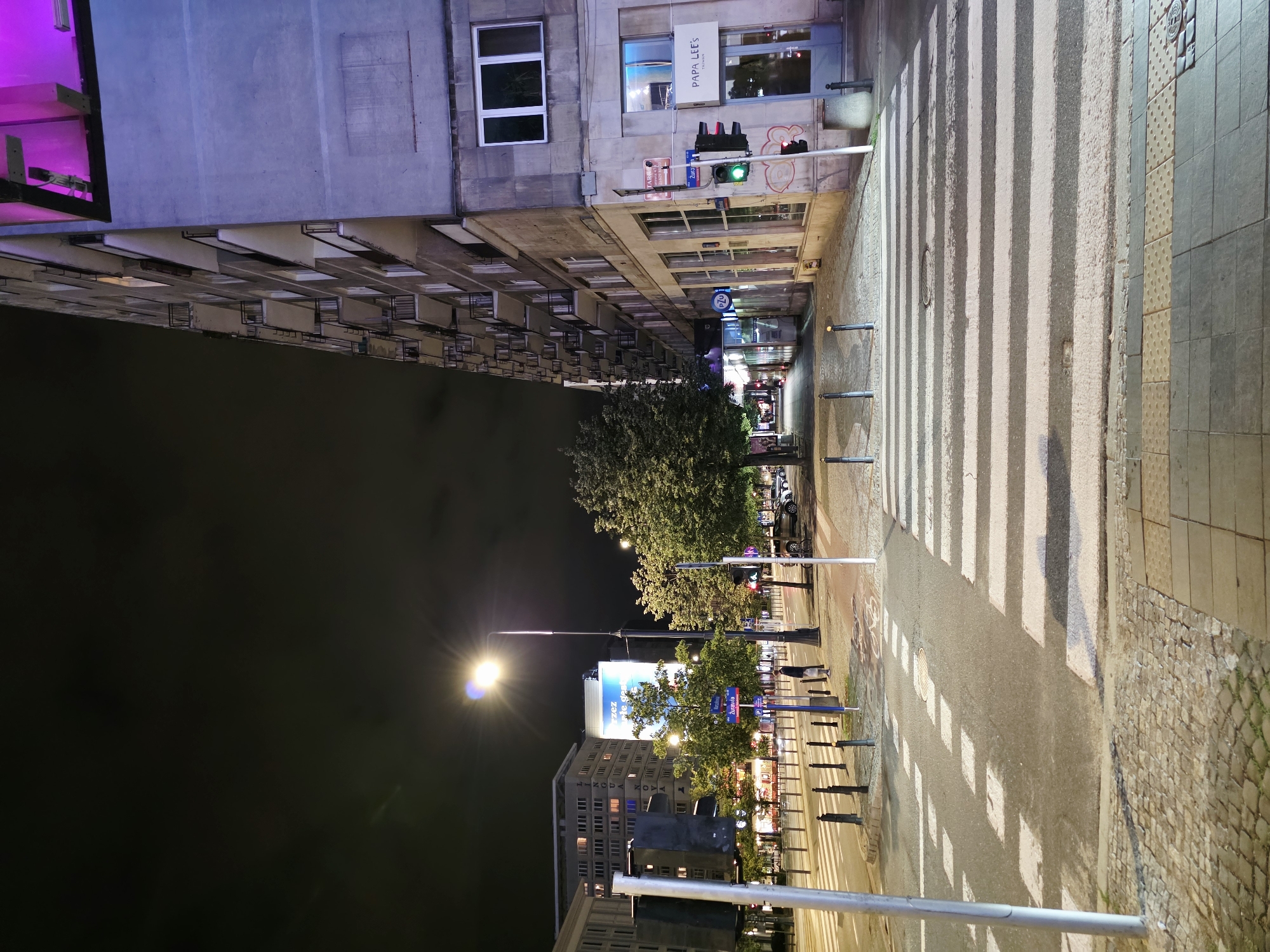 The image depicts a nighttime street scene in Warsaw, Poland, specifically at the intersection of Żurawia Street and Poznańska Street. The foreground features a pedestrian crosswalk with white stripes on asphalt. To the right, a modern building with several floors, characterized by balconies and windows, is visible. The building is partially illuminated by purple lighting on its upper floors and a sign for "PAPA LEE'S TAIWAN" is mounted near the entrance. A traffic light is showing a green signal. Trees line the street, and other buildings, including one with illuminated windows and a large digital billboard, are visible in the background. The street is illuminated by streetlights, and there are some cars parked along the side. A lone pedestrian is standing on the sidewalk near a lamppost, facing away from the camera. The sky is dark and cloudy.