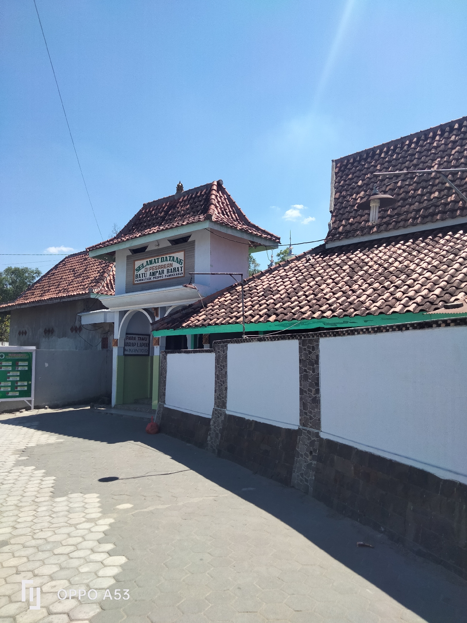 The image captures a building with traditional tiled roofs under a clear blue sky. The building appears to be a place of worship or a communal gathering spot, as indicated by a sign that reads "SELAMAT DATANG DI PESAREAN BATU AMPAR BARAT PANGEMBAK PUSPO PAMEKASAN". A second sign below it reads "PARA TAMU HARAP LAPOR KE KANTOR". The building is flanked by white walls with stone accents and a paved walkway in front. The location is identified as Badung, Indonesia. The scene is devoid of people, with only a shadow on the walkway suggesting a recent presence.