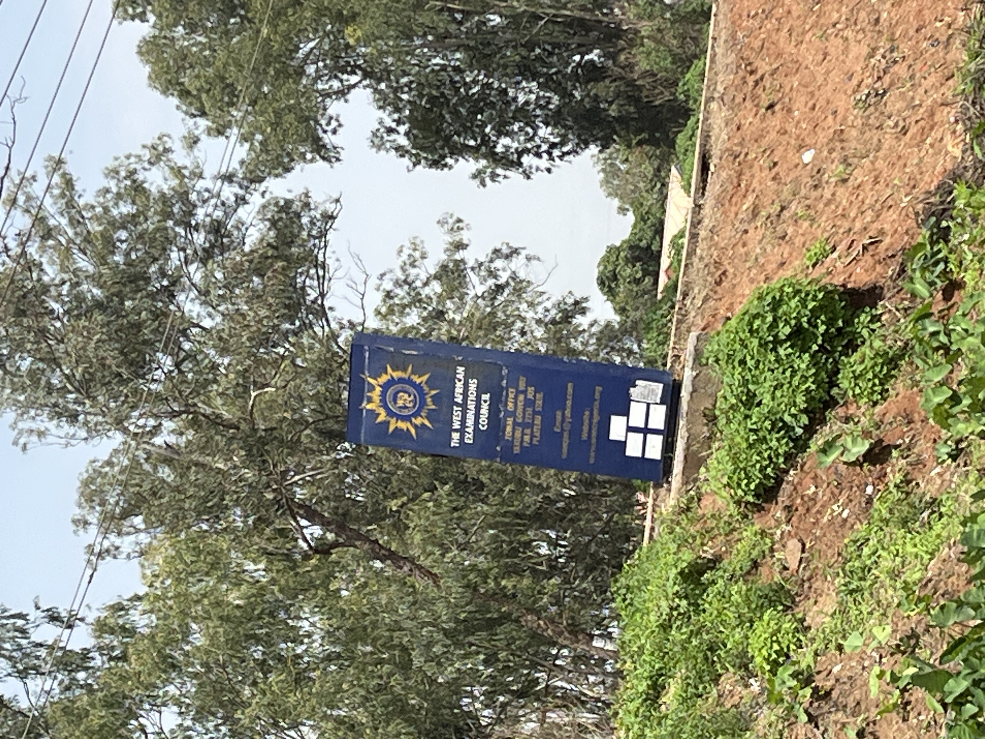 This media file is a vertical, medium-shot photograph taken outdoors during the day. The scene appears to be in Gura Top, Nigeria, as indicated by the location context. The primary object of focus is a blue sign for "THE WEST AFRICAN EXAMINATIONS COUNCIL". The sign is mounted on a pole and is partially obscured by trees. It features the council's emblem and text providing information about their ZONAL OFFICE in Plateau State, including an email address and website.
To the right of the sign, a dry, earthy hillside is visible, covered in patches of green vegetation. The background is dominated by lush green trees with a bright, overcast sky peeking through the branches. The lighting suggests it is daytime, possibly morning or afternoon, with diffused light due to the cloud cover. There are no people visible in the frame. The overall impression is one of a rural or semi-rural setting, likely near an administrative building or examination center. The photograph is taken from a slightly low angle, looking up towards the sign and the trees.