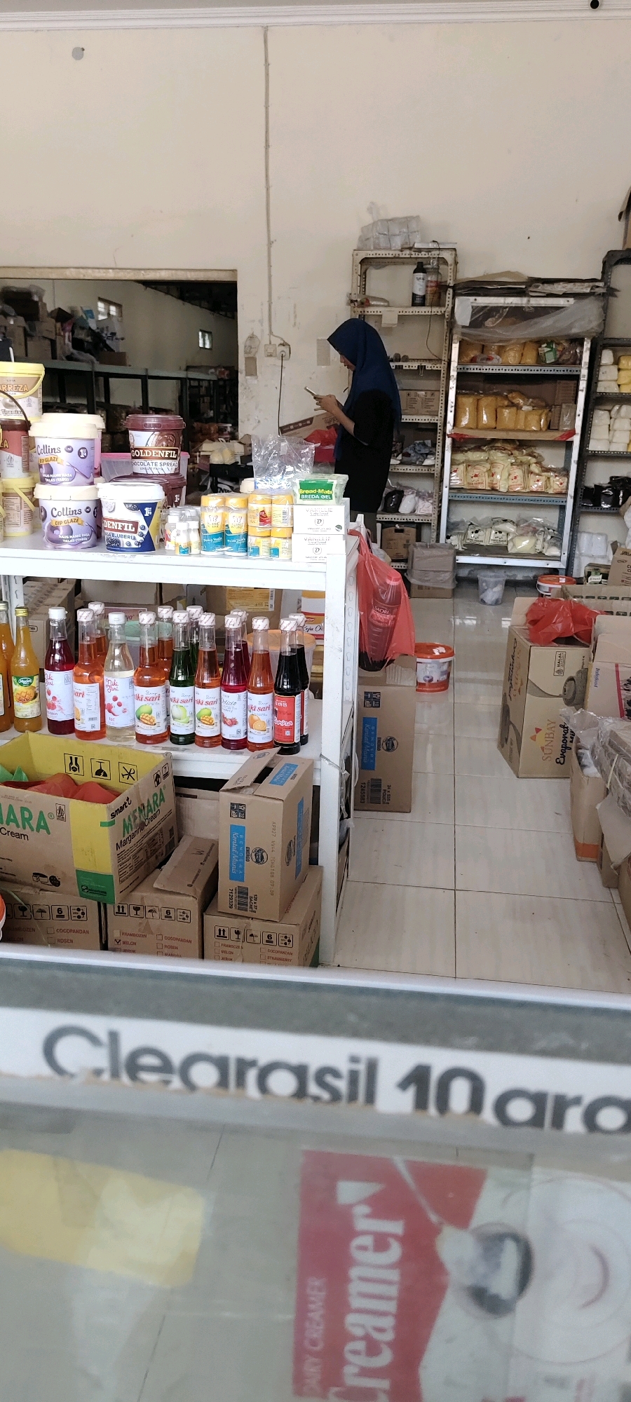 This is a medium shot, slightly angled, indoor photograph of a store, likely a grocery or convenience store, in Leteh, Indonesia. The store appears to be well-stocked with various food items, primarily packaged goods.
In the foreground, a large, rectangular glass display case is visible. A prominent white sign with black text is attached to the top of the display case, reading "Clearasil 10 ara". Inside the display case, a red and white carton of "Creamer" dairy creamer is partially visible.
The main focus of the image is a white shelving unit in the center of the store, laden with products. On the shelves, there are numerous bottles of colored liquids, likely syrups or juices, arranged in rows. Above these, various jars and containers of food products are stacked, including what appear to be chocolate spreads, glazes, and possibly other baking ingredients. Some of these containers have prominent branding, such as "Collins" and "Goldenfil".
To the right of the shelving unit, a woman wearing a dark blue hijab and dark clothing is standing and looking down at a mobile phone she is holding in her hands. Behind her, additional shelves are visible, filled with more packaged food items, including bags of yellow and beige powdered goods, and boxes.
The floor is tiled with light-colored square tiles. To the left of the shelving unit, several cardboard boxes are stacked on the floor. One larger yellow box has the text "Menara Margarine Cream" visible on it. Other brown cardboard boxes are also present, some with labels and shipping information.
The overall environment suggests a retail setting with a variety of goods for sale. The lighting is moderate, and the scene appears to be during daylight hours.