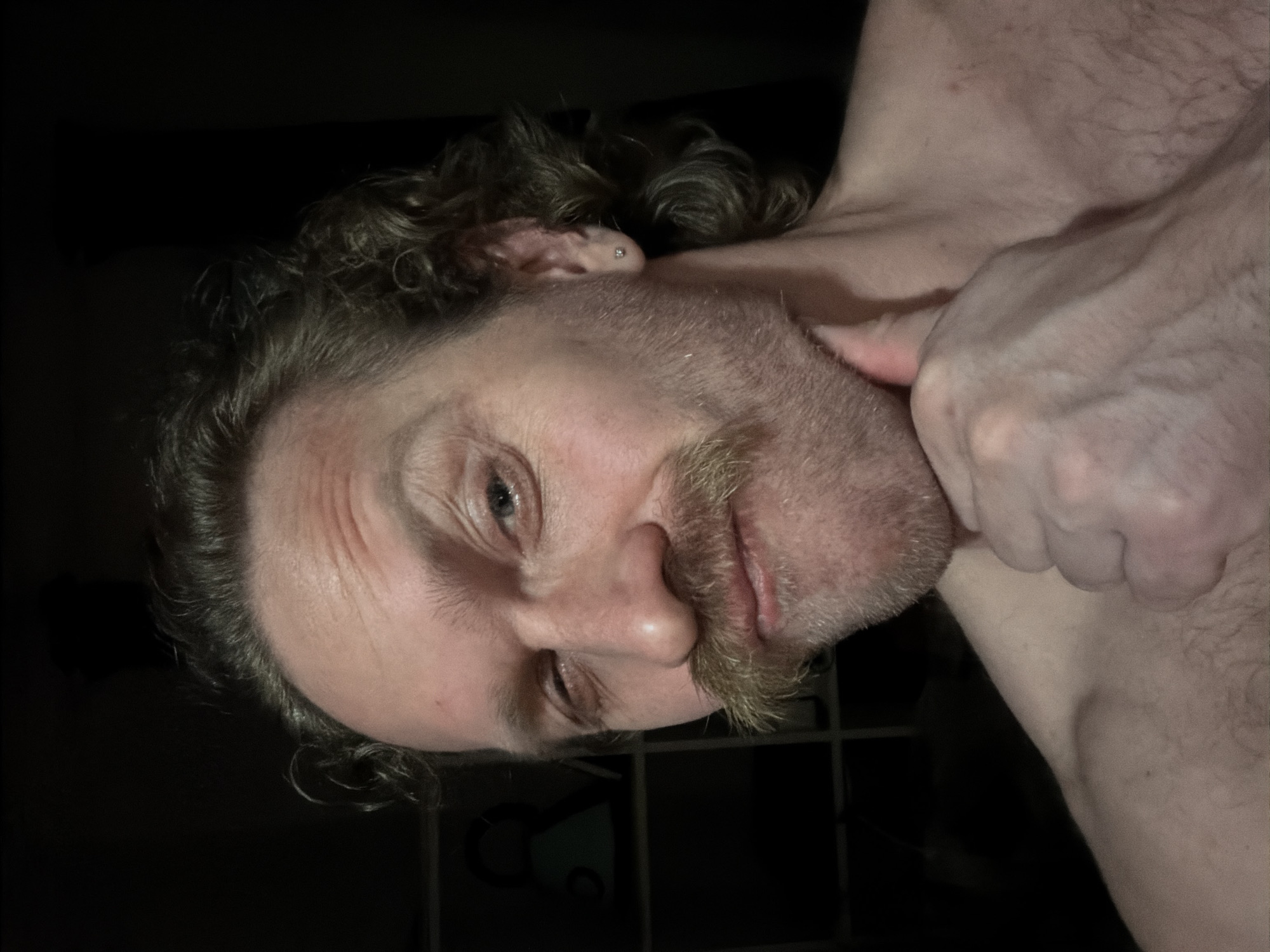 The media file displays a close-up, portrait-style image of a person, appearing male, situated in what seems to be a dimly lit indoor setting, potentially in Saint Petersburg, Russia. The individual's head is significantly tilted to the side, almost inverted, with their right eye looking directly towards the viewer. The expression on their face is ambiguous, possibly conveying intensity or contemplation, with visible wrinkles around the outer corner of the right eye. The left eye is partially closed or obscured.
The subject has curly, dark hair, a prominent mustache, and a beard. A small, silver stud piercing is visible on the helix of their left ear. Their skin appears fair, with visible body hair on the chest and shoulder area on the right side of the frame. The person's right hand is clenched into a fist and positioned near their neck or upper chest.
The background is predominantly dark and indistinct, offering no specific details about the environment other than its low light conditions. The lighting highlights features on the face and upper body, suggesting a direct or overhead light source. There is no visible text in the image.