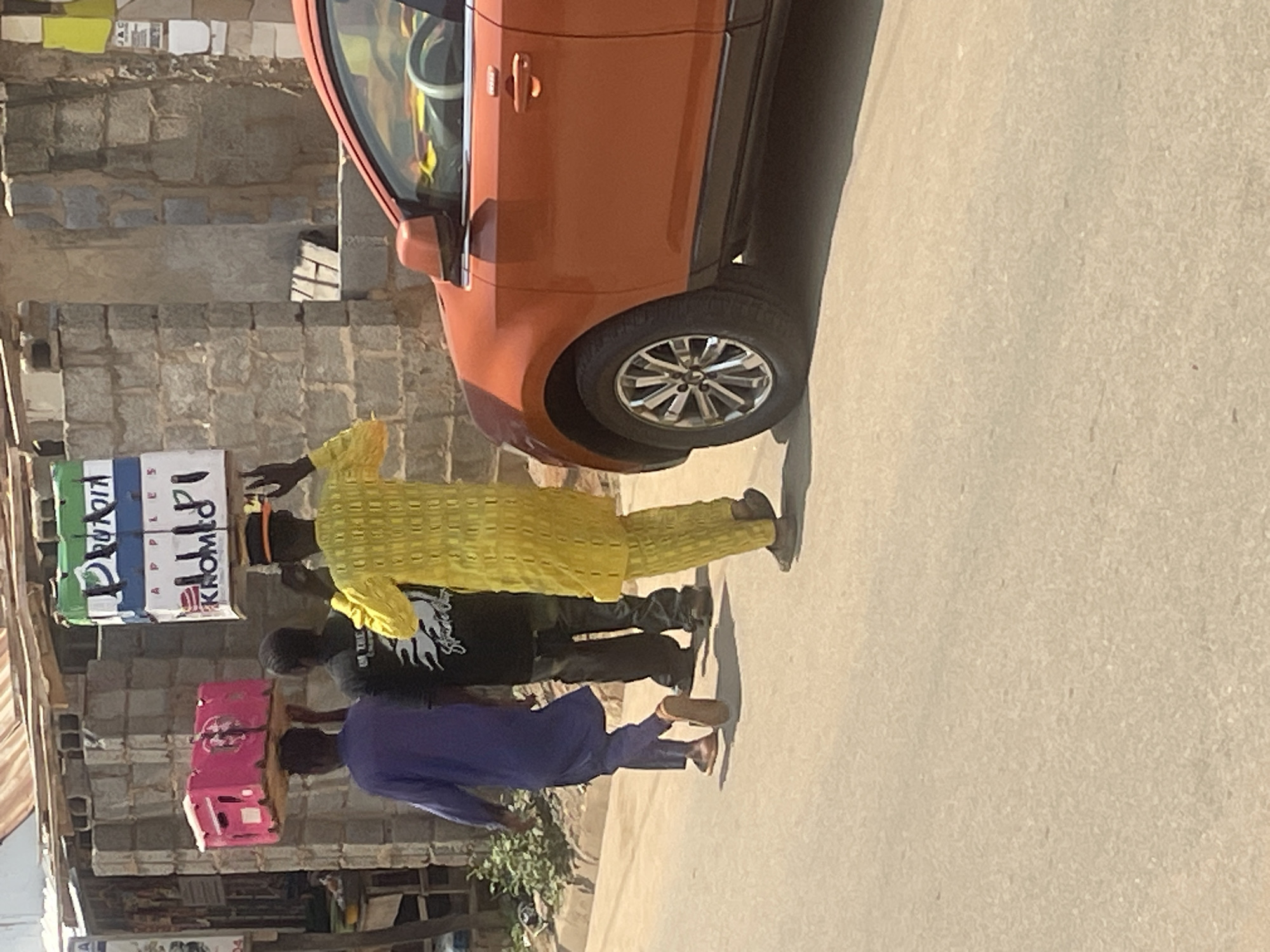 Three men are walking on a paved road in Abuja, Nigeria. The man on the left is wearing a long blue robe and walking away from the camera. He is carrying a pink box on his head. The man in the middle is wearing a black t-shirt with white writing and dark pants, and is also walking away from the camera. The man on the right is wearing a vibrant yellow outfit and a black and orange hat, and is walking towards the camera. He is carrying a large, multi-colored box on his head, which appears to contain food items. To the right of the men, the front section of an orange car is visible, parked on the side of the road. The background consists of a brick building with a corrugated metal roof, suggesting an urban or semi-urban environment. The road surface is rough and dusty.