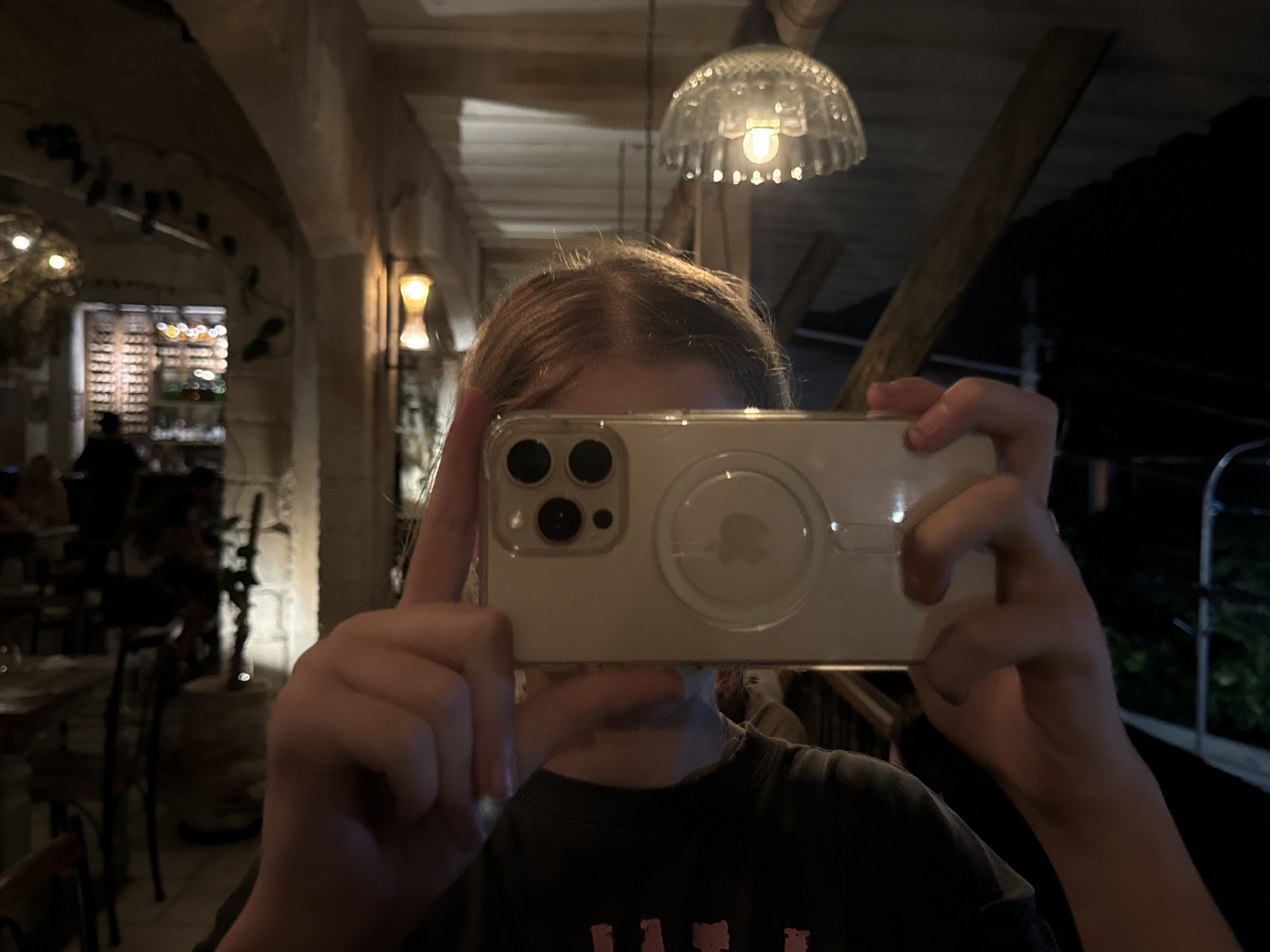 A close-up, dimly lit photo shows a person holding a white smartphone horizontally, obscuring their face. The person's forehead, hair (light brown/blonde), neck, and shoulders are visible. They are wearing a dark long-sleeved shirt with some faint, illegible text (possibly "TAT" or "CAT") on the front.
The smartphone is an iPhone model, visible by its distinctive camera module with multiple lenses and a flash. It is encased in a clear protective case, revealing the phone's back and a prominent circular MagSafe charging ring with a subtle Apple logo visible within it. The person's fingers are wrapped around the phone, with one thumb visible on the left side and other fingers on the right.
The background is a softly lit indoor space, possibly a restaurant or bar, characterized by arched stone or textured walls and wooden beams on the ceiling. Various light fixtures illuminate the scene, including an ornate glass pendant light directly above the person's head and a wall sconce further back on the right. In the left background, blurred tables and chairs are visible, along with what appears to be a wine rack or shelving system filled with bottles, indicating a dining or drinking establishment. The overall ambiance is warm and cozy due to the low lighting, suggesting it is nighttime. No specific weather details are discernible.