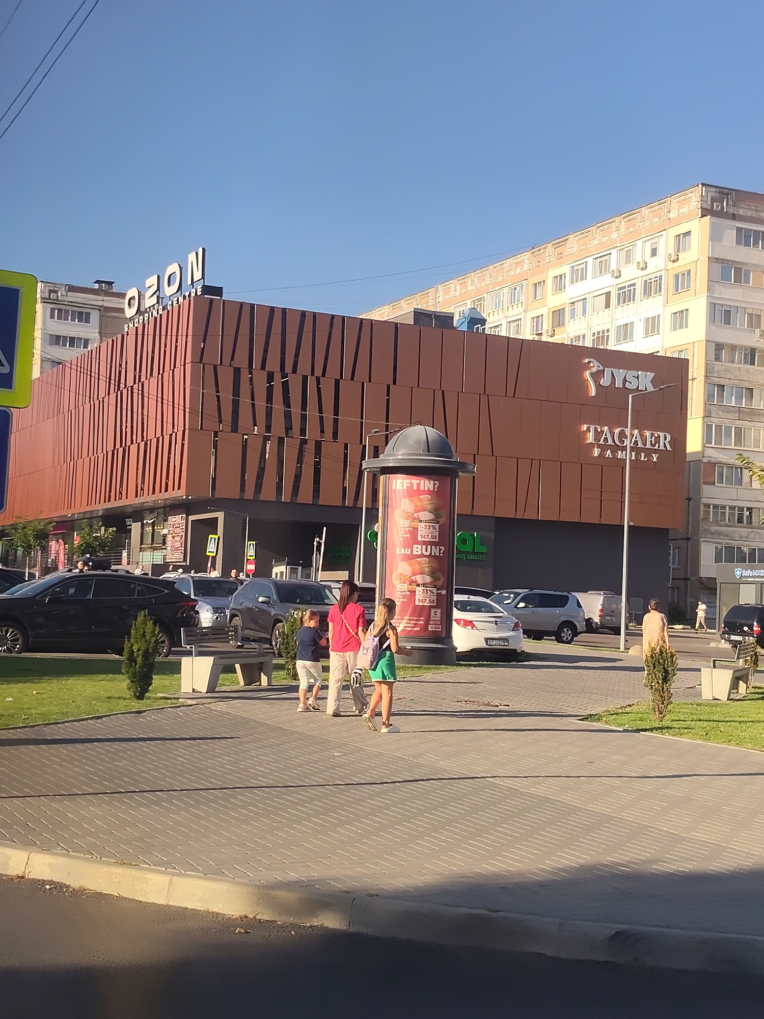 The image depicts the "OZON" shopping center, identified as being located in Chișinău, Moldova. The building features a distinctive brown facade with vertical, slatted elements. Prominently displayed on the building are the logos for "OZON SHOPPING CENTRE" and "TAGAER FAMILY," with "JYSK" also visible.
In the foreground, a paved pedestrian area is visible, with a street and sidewalk to the right. Several cars are parked in front of the shopping center, including a black sedan, silver SUVs, and a white sedan. A cylindrical advertisement pillar stands on the left side of the pedestrian area, displaying text and images, likely promoting discounts.
Several individuals are present. Two young girls are walking on the paved area towards the right side of the frame. A woman in a pink shirt and white shorts is walking with them. Further to the right, a woman wearing a light-colored dress is walking away from the camera. In the background, near the shopping center entrance, several other individuals are visible, including a person wearing a yellow vest, suggesting they might be an employee or security personnel.
The environment appears to be an urban setting, characterized by modern commercial architecture and public spaces. The sky is clear and blue, indicating daytime. Green grass and small trees are visible in landscaped areas around the shopping center and pedestrian walkway. A traffic light pole with a blue sign is also present on the far left of the image.