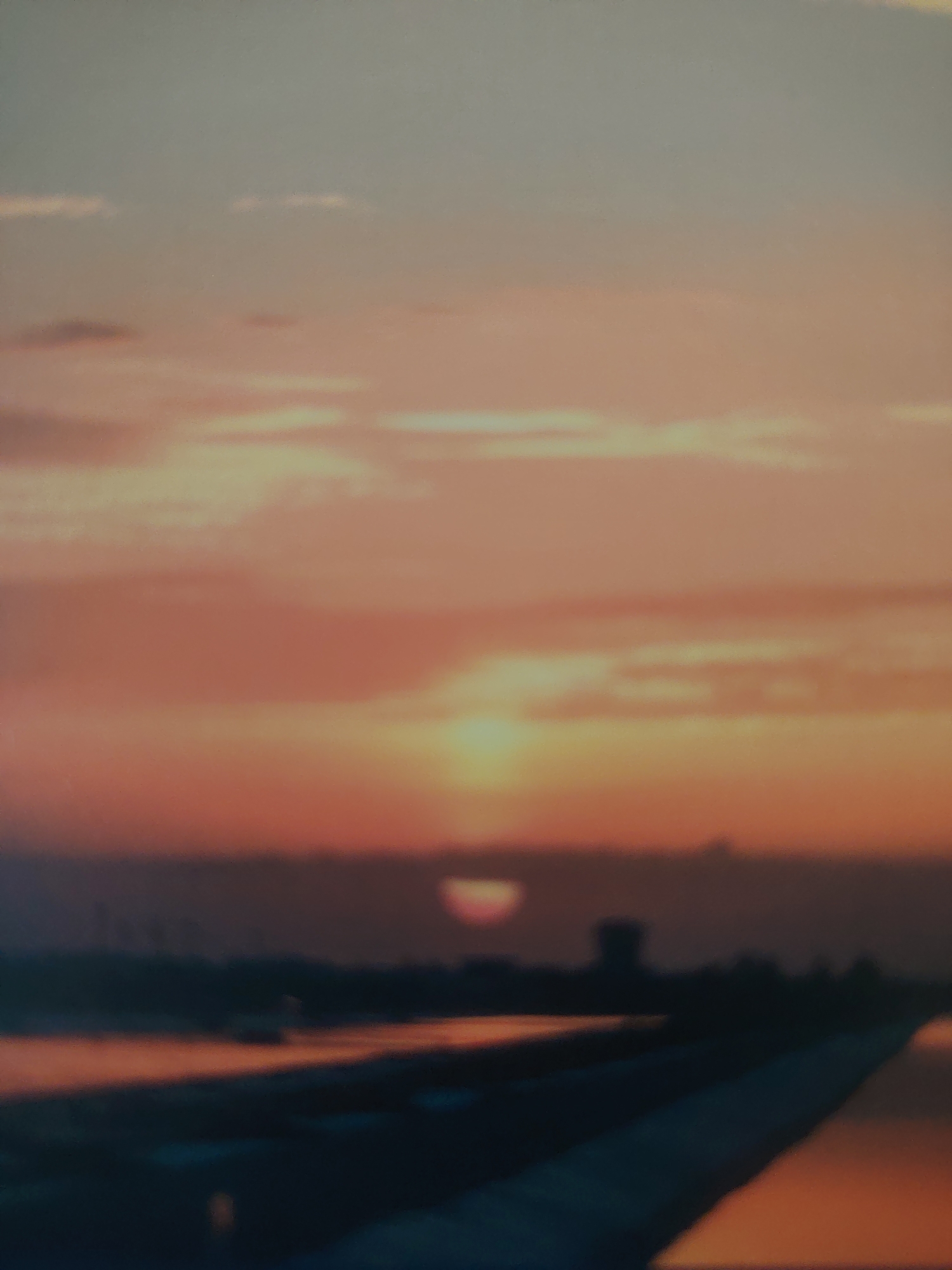 The image captures a hazy sunset over a body of water, likely a canal or river, with a distant shoreline visible. The sky is dominated by warm hues of orange, pink, and pale blue, with soft clouds. A dim, partially obscured sun is setting near the horizon, casting a reddish glow. In the middle ground, a dark silhouette of what appears to be a building or structure, possibly a tower, stands against the sky. The foreground shows a dark, indistinct area that could be land or a path, with reflections of the sunset on the water's surface. The scene suggests a waterfront area within a city, but the specific location in Bahshitpa, Uzbekistan, cannot be determined from the visual information alone. There are no visible people or clear actions taking place.