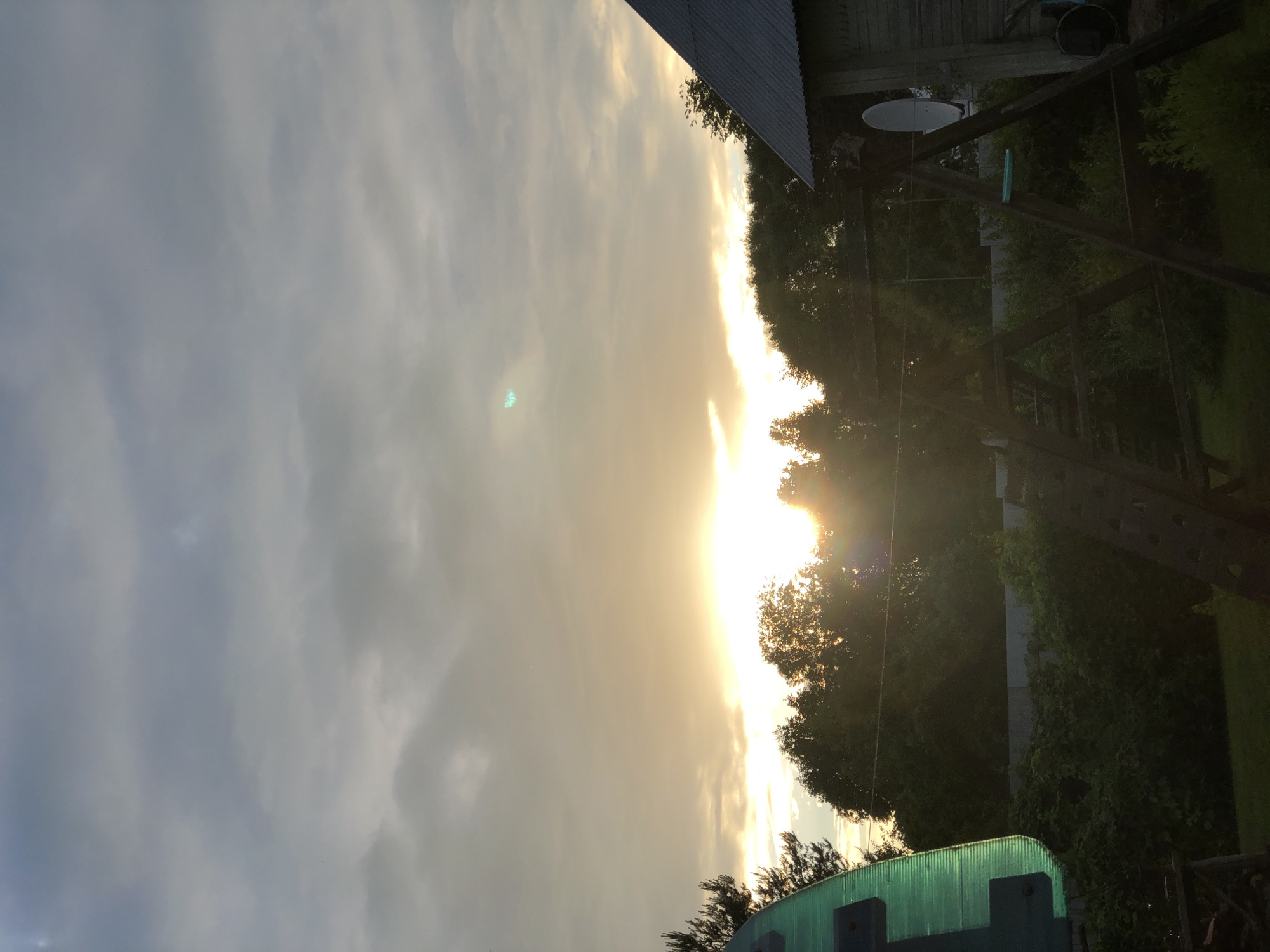 The image captures a natural landscape in the city of Zhukovo, Russia, during what appears to be sunset or sunrise. The sky is filled with overcast clouds, with a bright area of sunlight breaking through the clouds near the horizon, casting a warm glow. Silhouetted against the sky are the tops of lush green trees, suggesting a rural or park-like setting. In the foreground, elements of a backyard or recreational area are visible. On the left side, a large, curved, translucent green object, possibly part of a play structure or a garden feature, is partially visible. In the center and right, a wooden swing set or similar play equipment stands, with its dark wooden frame clearly defined against the background. To the far right, a rustic wooden structure with a corrugated metal roof, possibly a shed or outbuilding, is present, with a white satellite dish mounted on its side. The overall scene is one of a tranquil, outdoor environment with a focus on natural light and the integration of man-made recreational elements into the landscape.