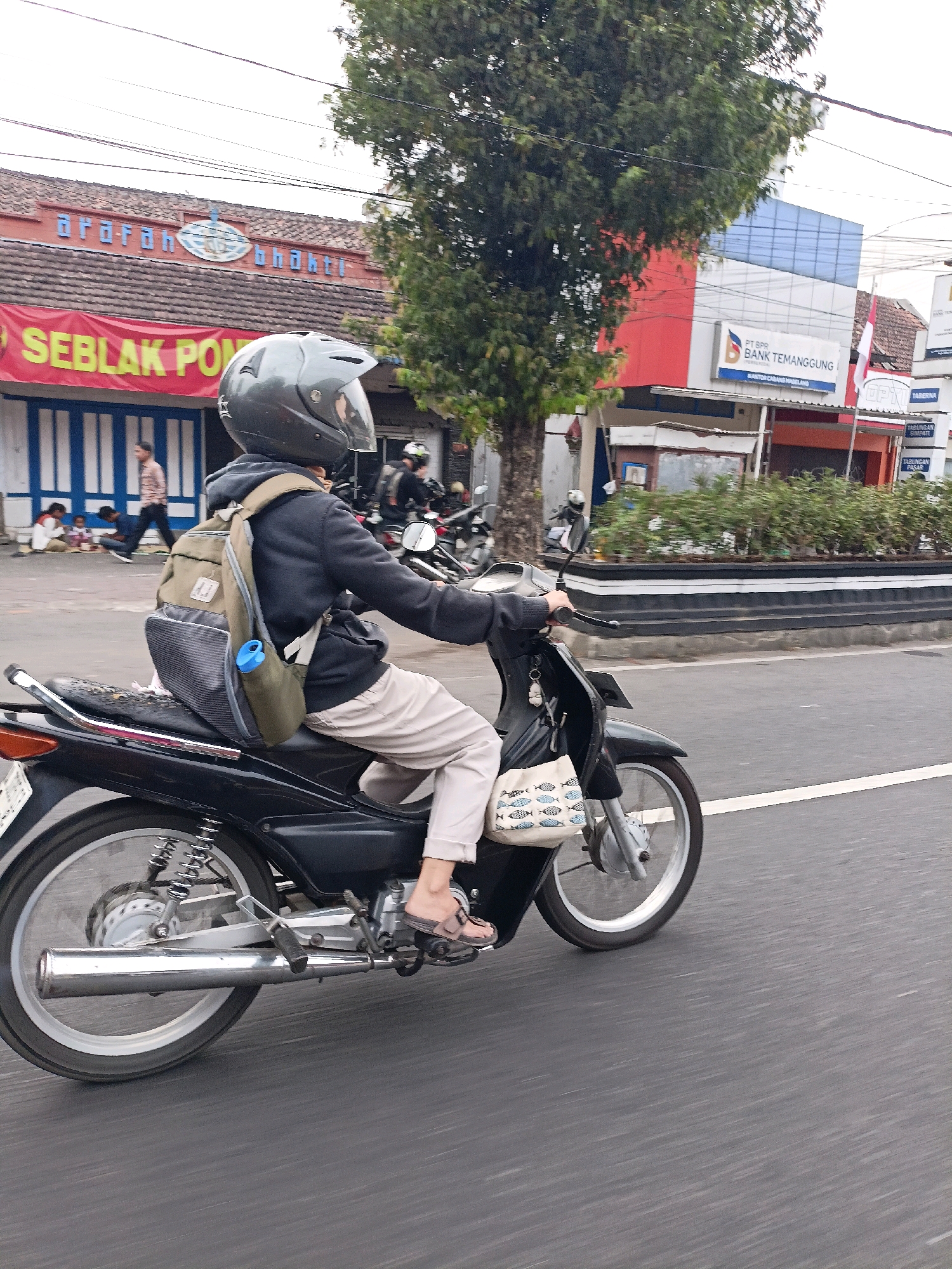 A person wearing a helmet and backpack rides a black motorcycle in motion on a street in Magelang, Indonesia. The person is wearing light-colored pants and sandals, and a fabric bag with a fish pattern is attached to the front of the motorcycle. In the background, buildings and trees are visible. One building has signage that reads "ararah bhakti" in blue letters and a red banner with the text "SEBLAK PO" in yellow. Another building displays "PT BPR BANK TEMANGGUNG". The road is paved and has a white line marking. The image is taken from a side angle, capturing the motorcycle and rider in motion.