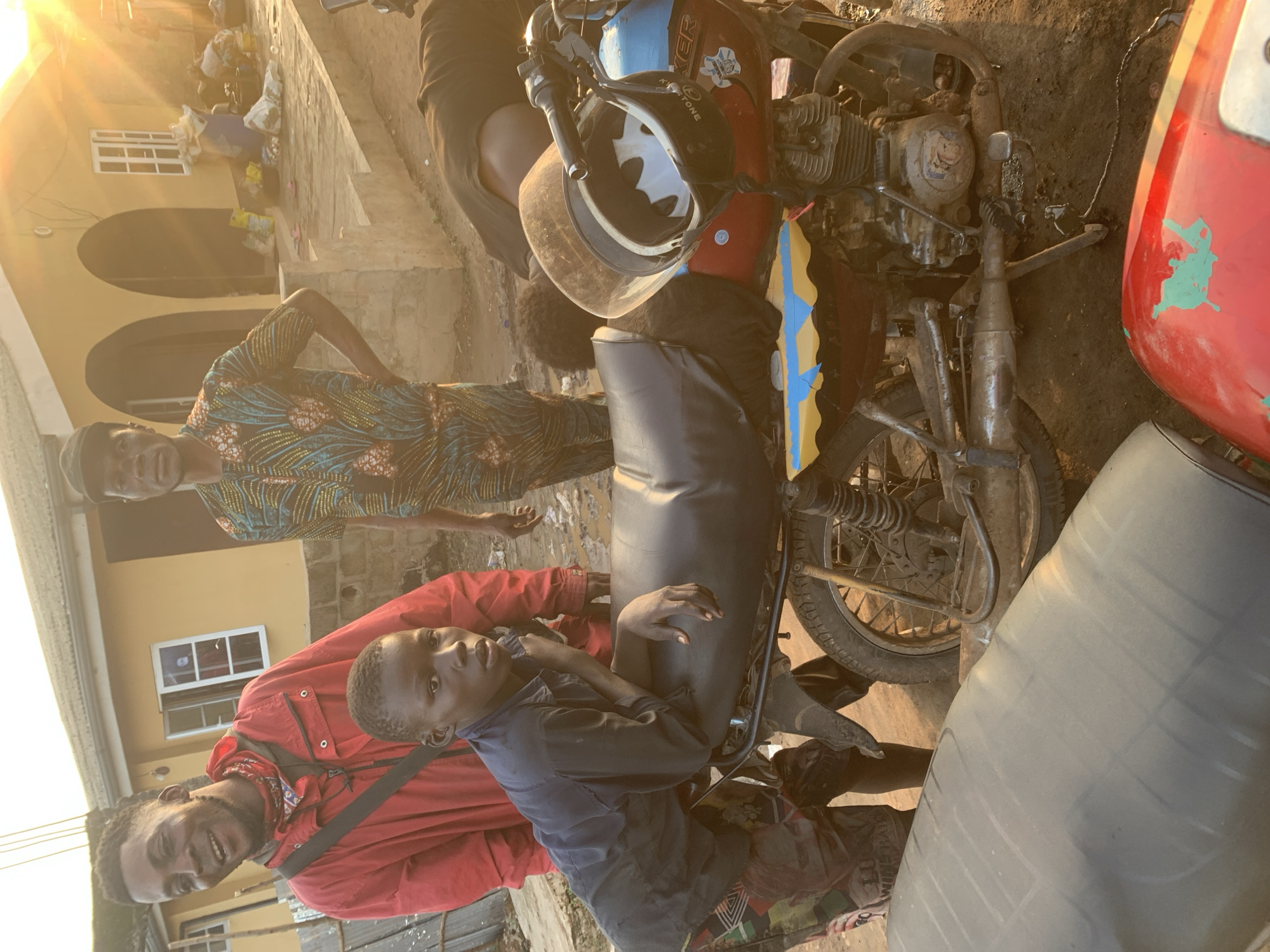 The photograph depicts a scene in the city of Shagamu, Nigeria, during what appears to be a sunny day, possibly late afternoon due to the warm lighting. In the foreground, a young boy, appearing to be around ten years old, is leaning on a motorcycle. He is wearing a dark blue long-sleeved shirt and patterned shorts. The motorcycle is a standard commuter model, painted red, blue, and yellow, and appears somewhat weathered. A helmet with a distinctive bat symbol on it rests on the motorcycle's seat.
Behind the boy, two adults are visible. To the left, a man in a bright red jacket and a patterned bandana around his neck is looking towards the camera with a wide smile. He has a sling bag across his chest. To his right, another man is standing, wearing a patterned shirt and a dark cap. His expression is neutral as he looks forward. Further in the background, a building with a yellow facade and arched doorways can be seen. Piles of items, possibly household goods or merchandise, are scattered on the ground near the building. The ground appears to be unpaved, with patches of dirt and possibly mud. Another motorcycle, largely out of focus and covered in a grey material, is visible in the lower left portion of the image. The overall atmosphere suggests a casual, possibly public, gathering in a residential or commercial area of Shagamu.